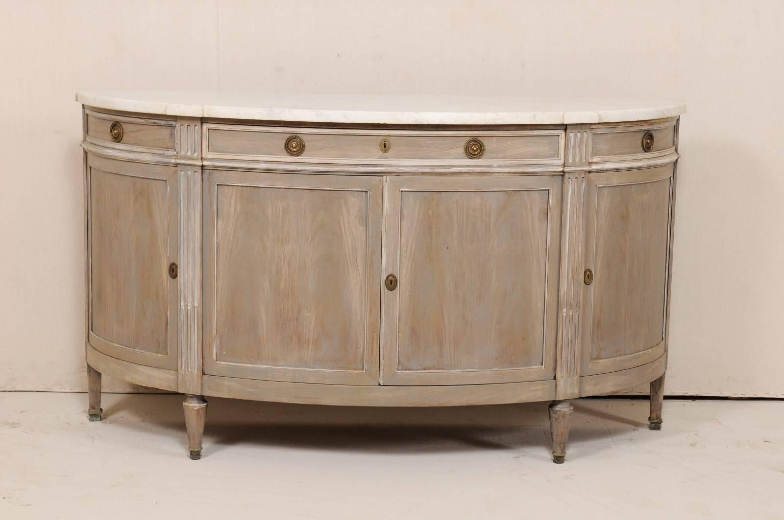 An French marble-top demilune cabinet from the early 20th century. This antique French cabinet features an elegantly curved demilune body, with fluted details down each of the four side posts, topped with white marble and raised on pegged feet.