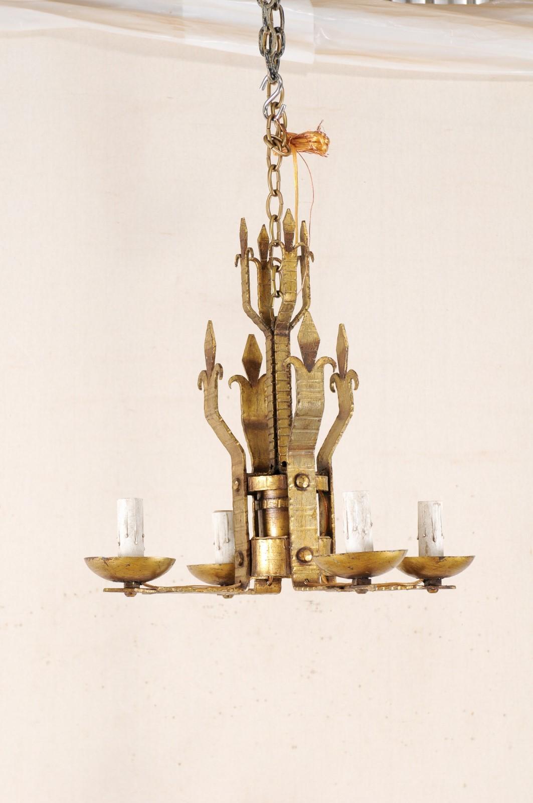 A French four-light gold colored iron chandelier from the mid-20th century. This vintage light fixture from France features a stylized Fleur de Lys body with four arms extending straight out from bottom, each supporting a cupped iron bobèche and