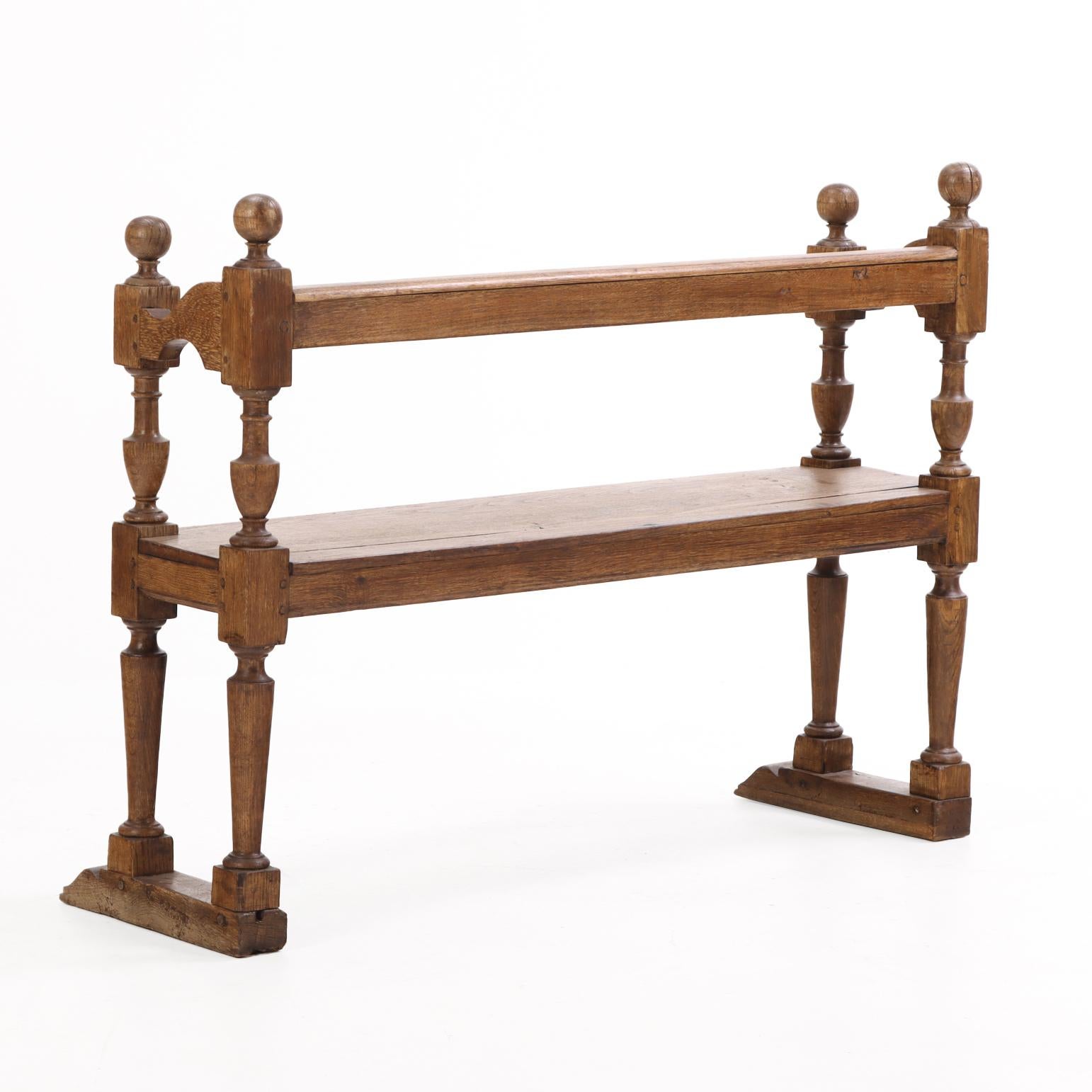 The design features a singular straight back rail set above a wide plank seat, all supported by bold turned legs surmounted by ball finials and held together at base with with two shoe feet. The combination of robust joinery and sculptural turning