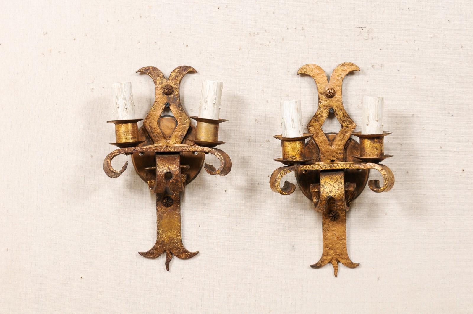 A French pair of two-light iron sconces from the mid-20th century. This vintage pair of French sconces have a linear back plate with splayed top, pierced diamond cutout at center, and ending with a stylized fleur-de-lys motif. A horizontally placed