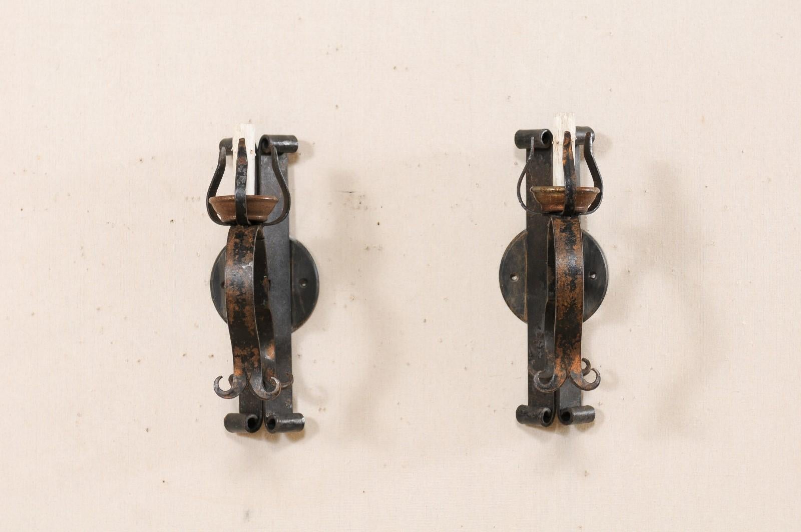 A French pair of iron sconces from the mid-20th century. This vintage pair of sconces from France each feature a vertical V-mount, raised back plate, with each tip terminating into a curl, and a single light which rests atop the arm, which is nicely