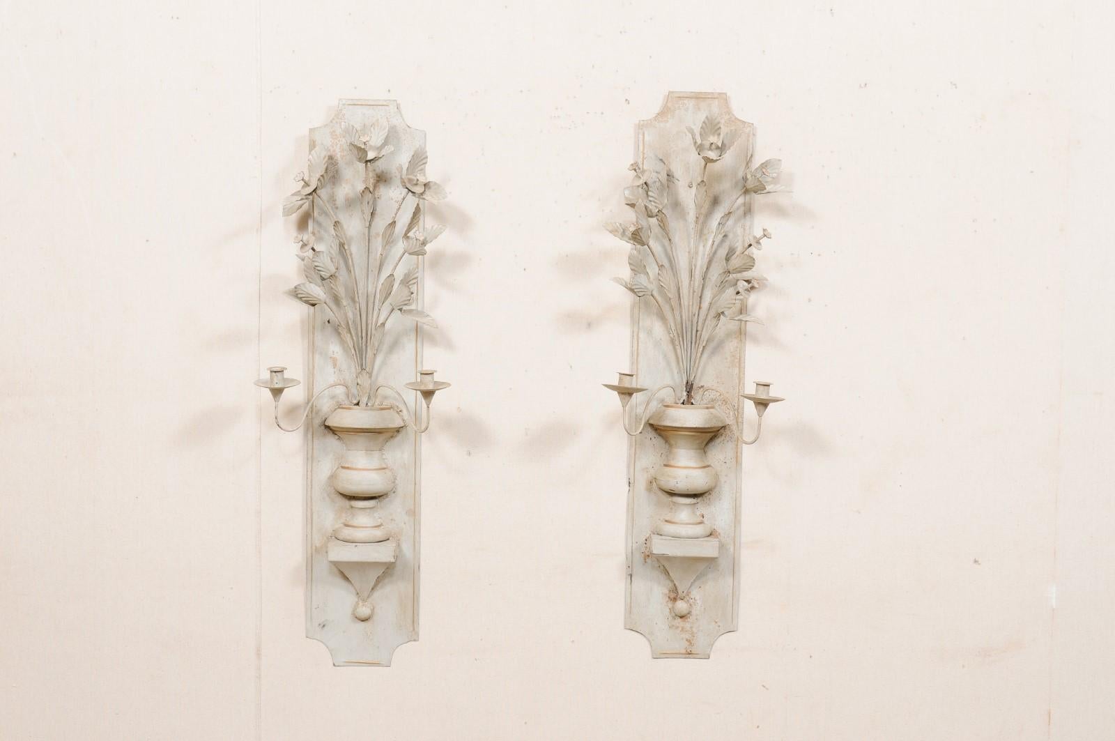 A pair of French two-light sculpted and painted metal candle wall sconces from the mid 20th century. This vintage pair of decorative wall sconces from France feature elongated metal backplates with sculpted urns with a bouquet of flowers splaying