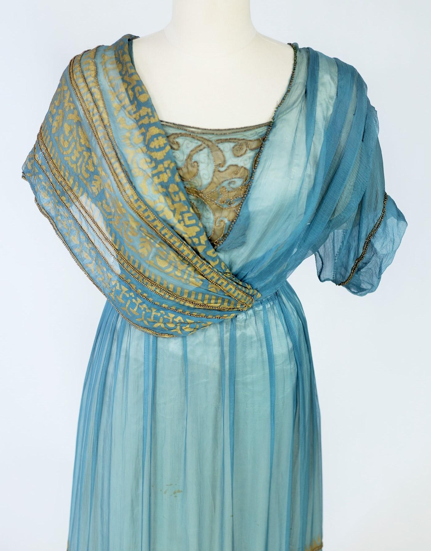 Circa 1910-1915
France
Evening dress in blue silk chiffon printed with geometric friezes with embroidery of golden rectilinear beads from a fashion house in Lyon. High-waisted cut with asymmetrical bustier by a draped shawl effect crossed on the