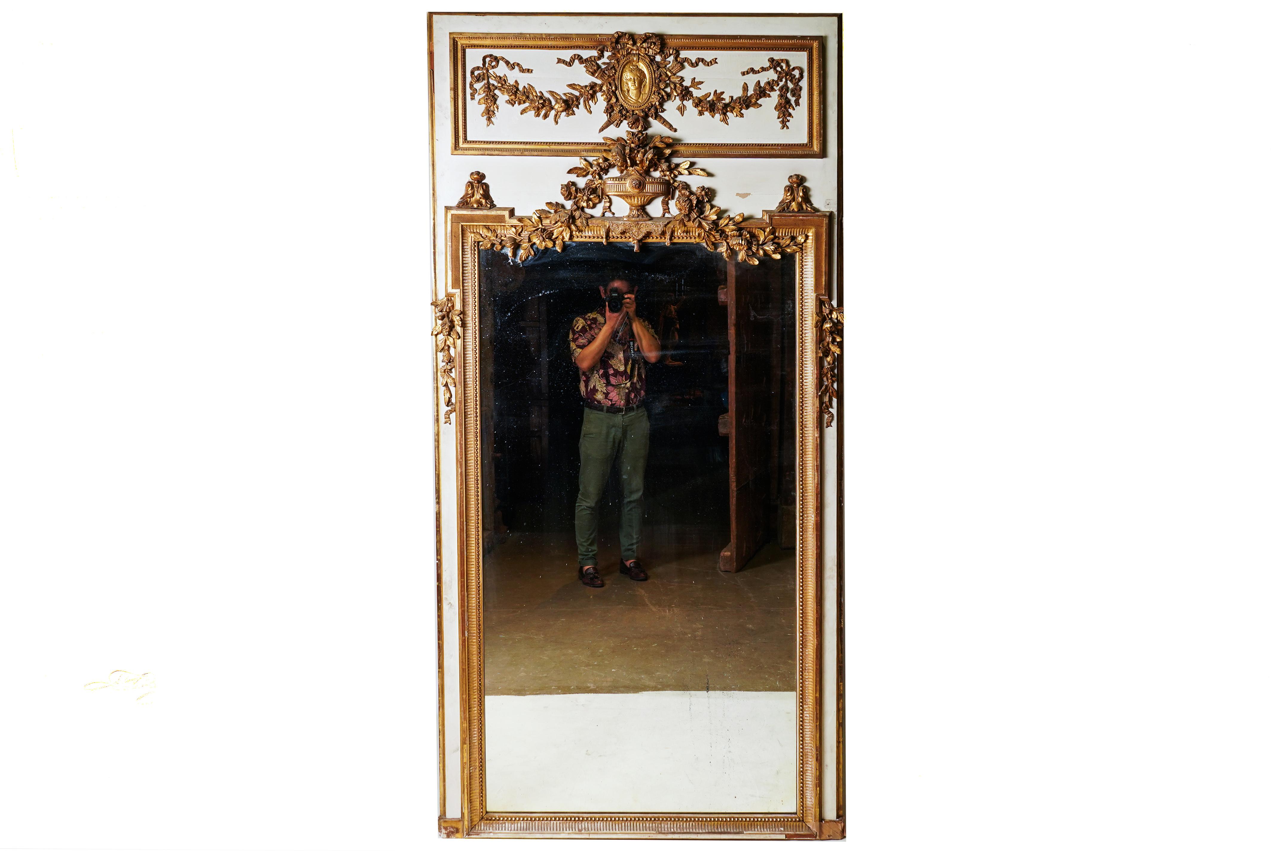 This impressively scaled and richly decorated trumeau mirror is from Paris, France and made from pine wood, gesso and genuine gold gilt. The mirror glass is antique glass with silvered backing, possibly original to the piece. The mirror has minor