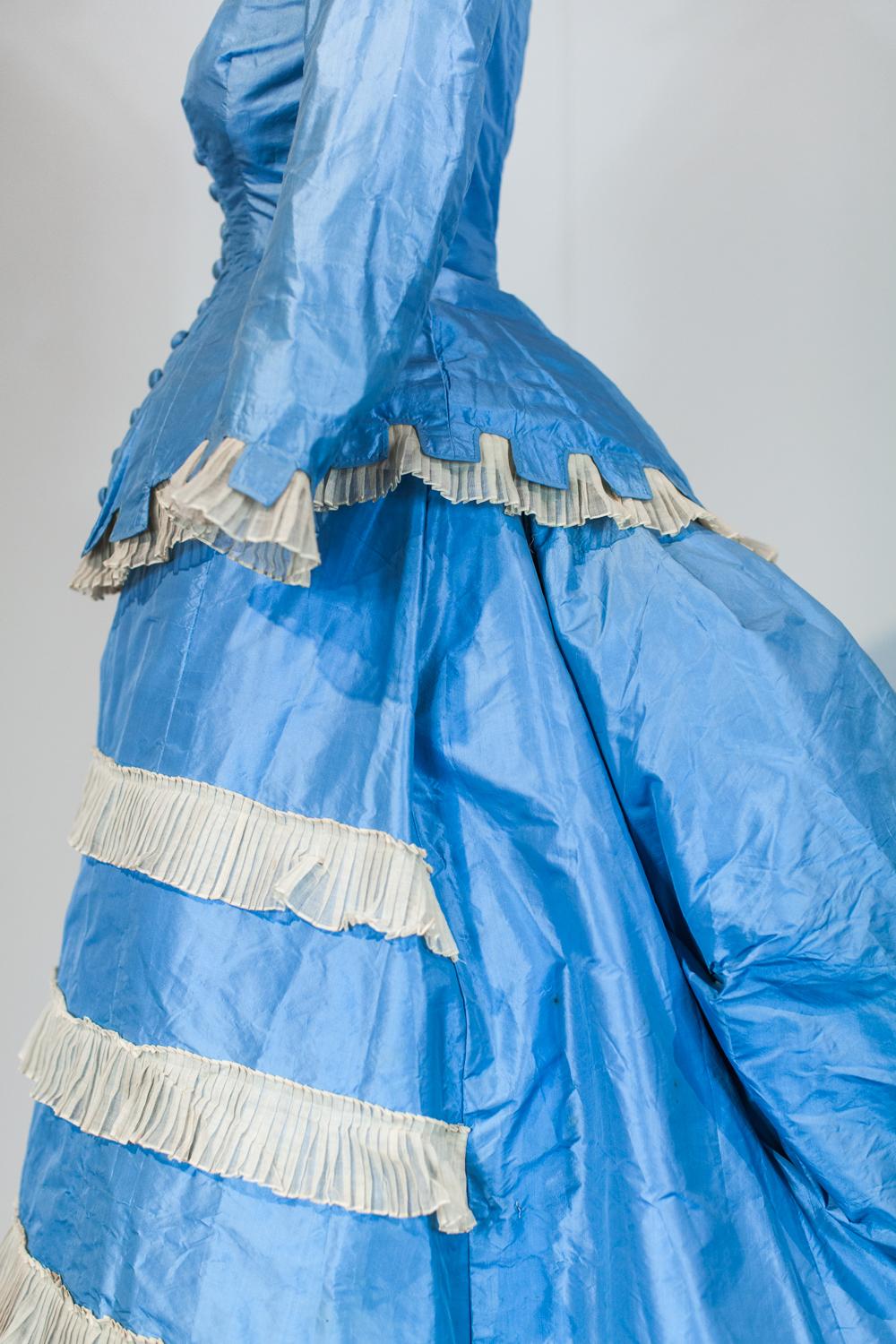 Circa 1870/1875
France.

Day dress with  princess, two-part bodice and skirt on a bustle cage. Sky-blue taffeta decorated with starched cream gauze called tarlatane, finely wrinkled and crenellated à la grecque. Boned bodice with long basque closing