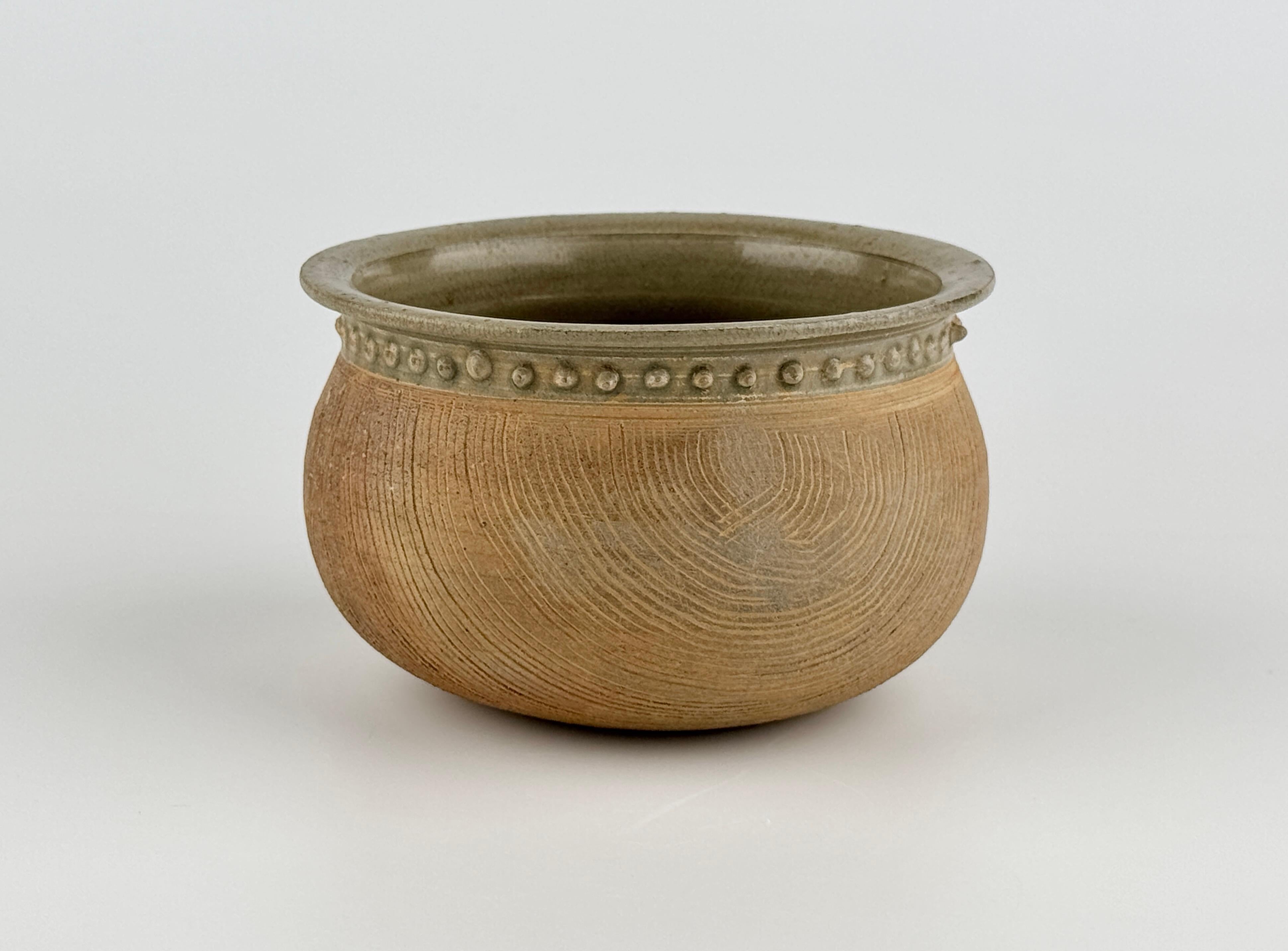 Este recipiente presenta un cuerpo suavemente redondeado con un borde grueso y acampanado, mientras que la parte inferior exterior está decorada con líneas incisas uniformemente talladas, inspiradas en la cestería. Una banda continua de pequeñas