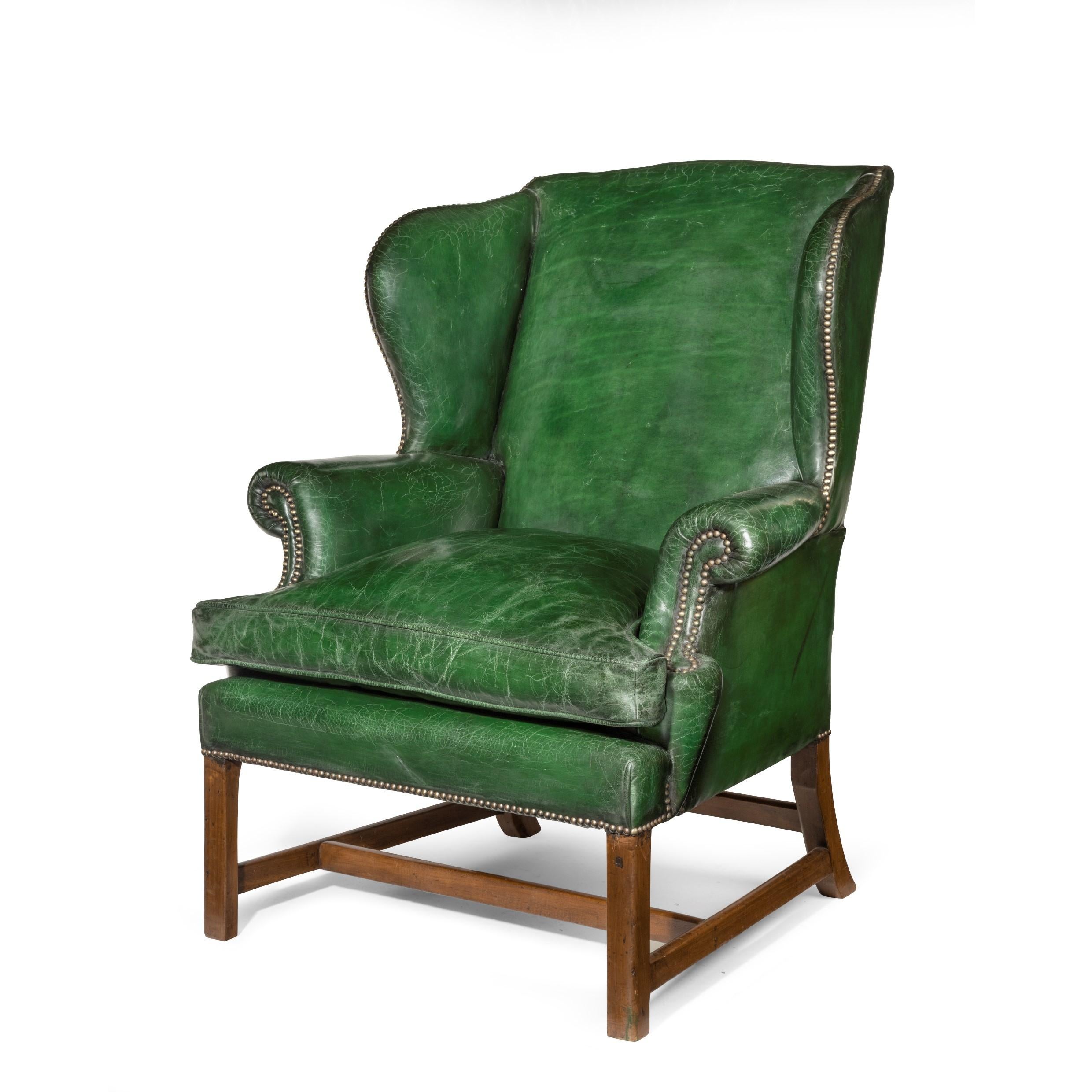 A generous George III mahogany wing armchair, of typical form with wings and scrolling arms, raised on square chamfered front legs and swept chamfered back legs, reupholstered in distressed green leather with brass studs, replaced castors. English,