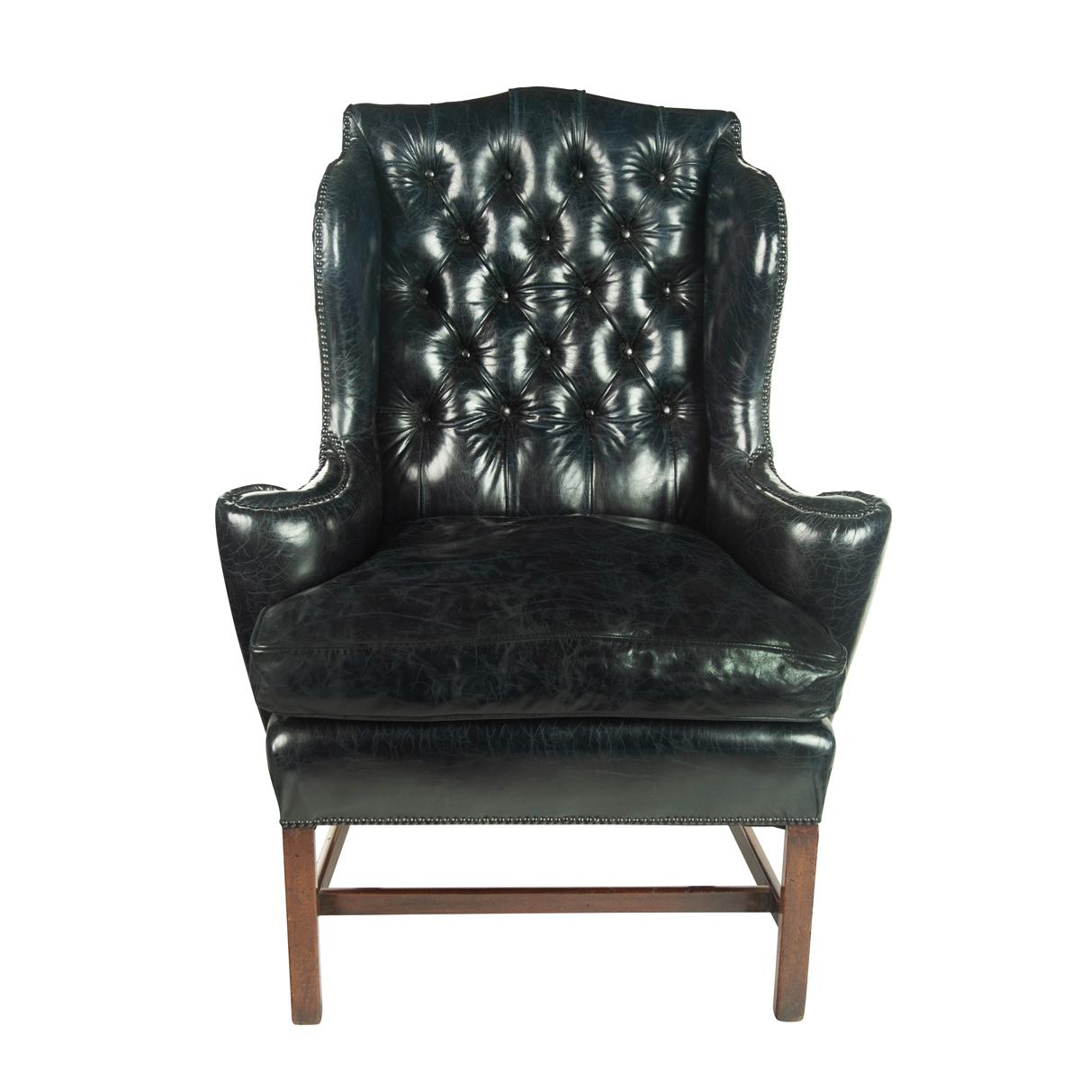 A generous George III wing arm chair, the shaped deep-buttoned back with scrolling arms, reupholstered in distressed blue-black leather, raised on four square section legs joined by an H shaped stretcher. English, circa 1800.
  