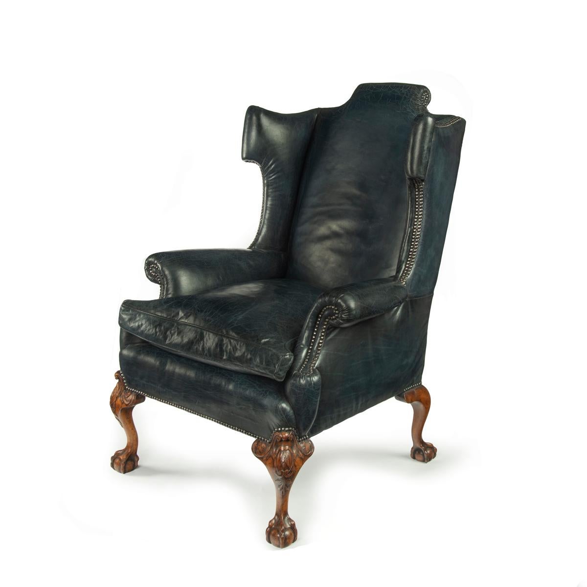 A generous late Victorian walnut wing arm chair, in the Georgian style, with scrolling wings, shaped top rail, carved on the front knees with confronting C-scrolls and foliage, all four legs with claw and ball feet, reupholstered in distressed blue