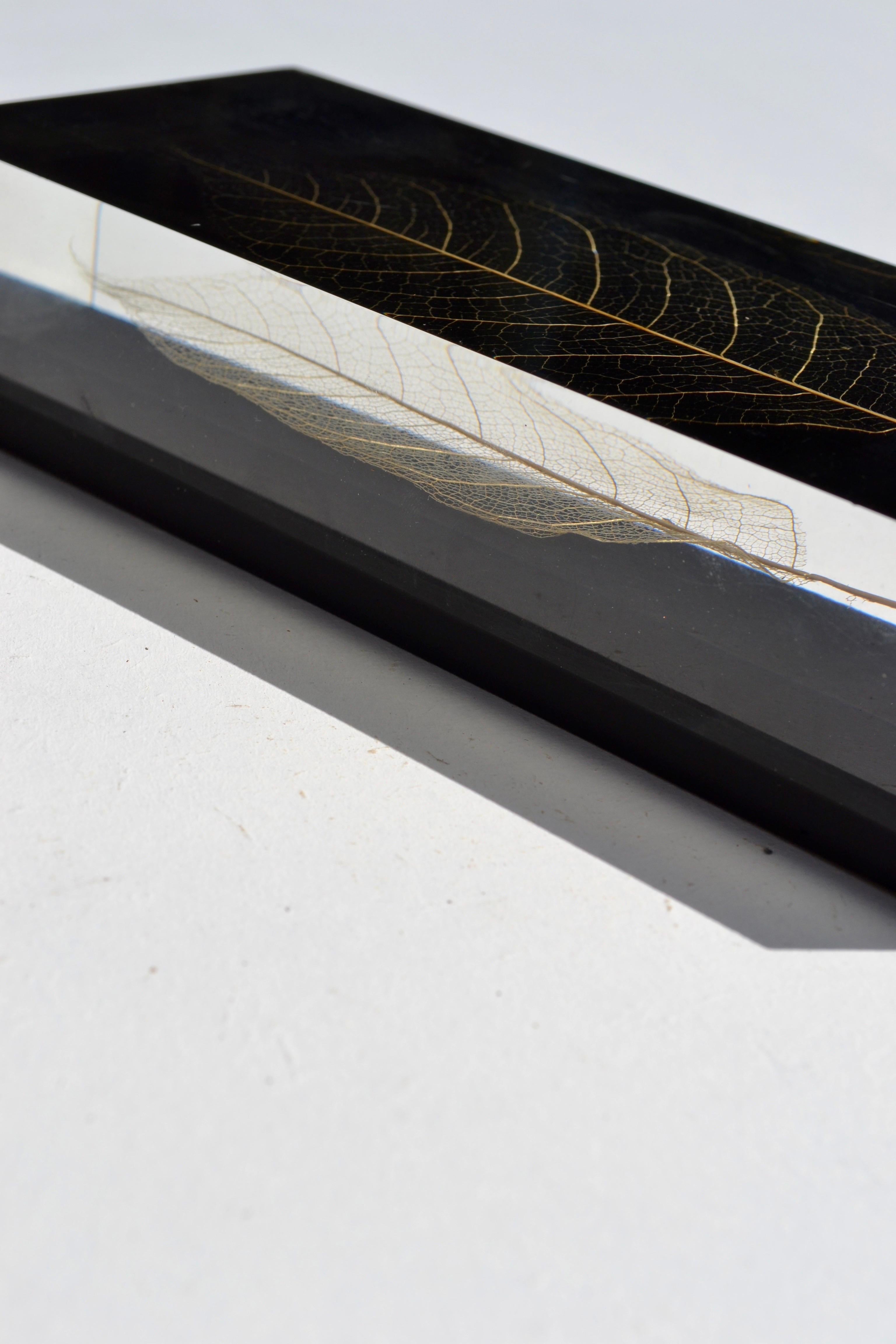 
A striking rectangular resin paperweight from the 1970s, featuring a delicate skeleton leaf, gilded before being suspended in layers of black and translucent resin. The minimalist, monolithic form—perfectly parallelepiped—gives the object a