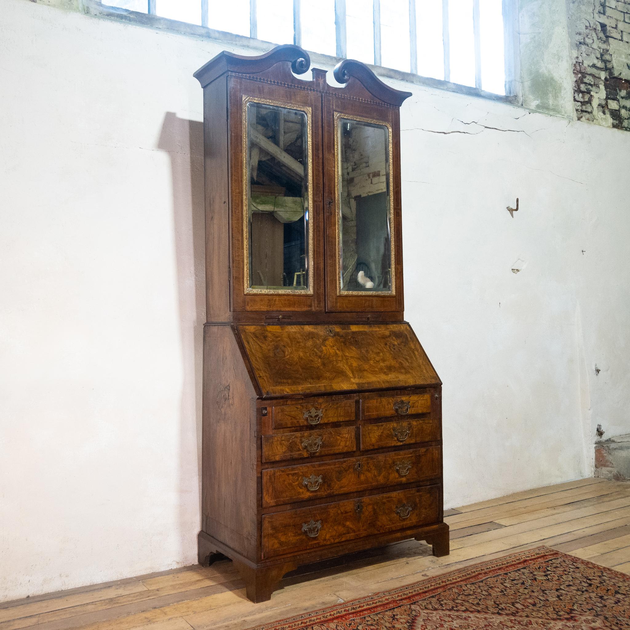 Una superba libreria bureau di Giorgio I in noce figurato e con fasce di piume. Mostra un affascinante frontone a collo di cigno, di sezione sagomata a cavetto, con dettagli in ebano. Con una coppia di ante a specchio al mercurio, incorniciate da un