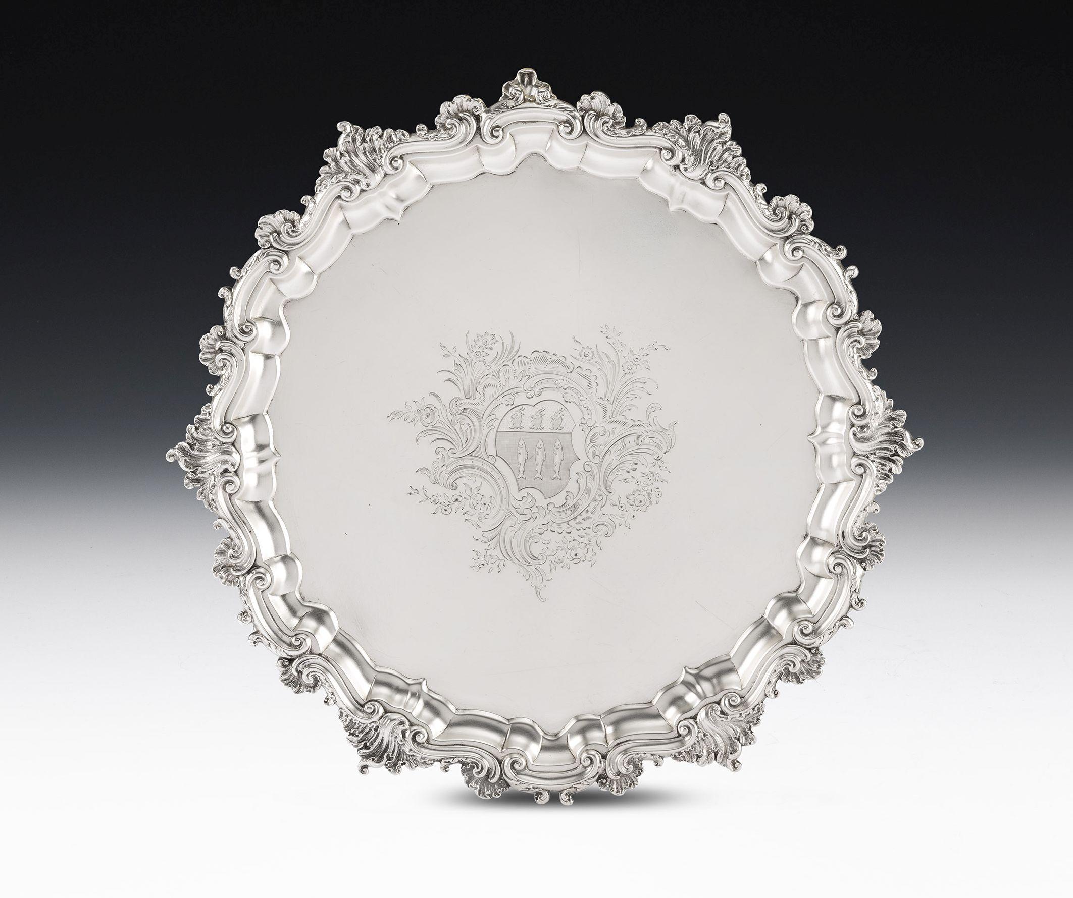 The Salver stands on three substantial raying shell feet with leaf capped scroll legs above. The main body has a raised shaped scroll rim with cast Rococo shells attached, flanked by foliate motifs. The centre is beautifully engraved with a
