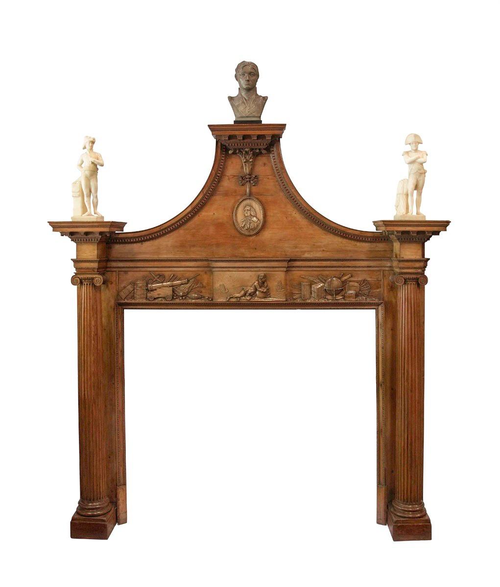 This pine chimneypiece has a swan-neck pediment supporting three pedestals, with dentil and leaf borders.  There is a central limewood oval portrait of Robert Marsham, 2nd Baron Romney carved in relief and suspended by acanthus and tied ribbons. 