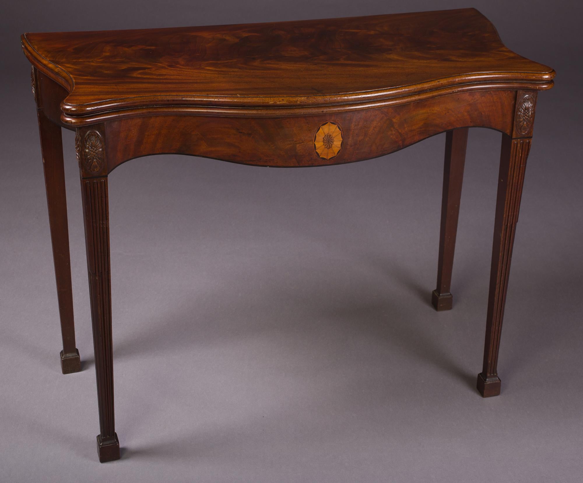 A George III Chippendale period Mahogany card table.
The well figured serpentine top with a moulded edge,
above a veneered frieze, with carved mounts a central satinwood sun burst,
resting on square tapered fluted legs with block feet.