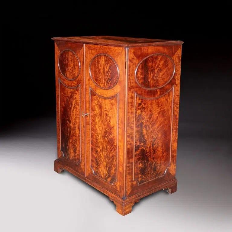 A handsome flame mahogany collector’s cabinet, circa 1800, the moulded rectangular top above a pair of oval and shaped panelled cupboard doors, the inset veneers being within raised borders of ‘woven’ boxwood and ebony veneered and with strung inlay