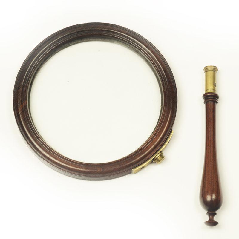 A George III gallery magnifying glass, the circular glass within a mahogany frame, the baluster handle screwing into a brass bracket in the rim, the handle with a brass collar. English, circa 1800.

Footnote: The gallery glass is a type of