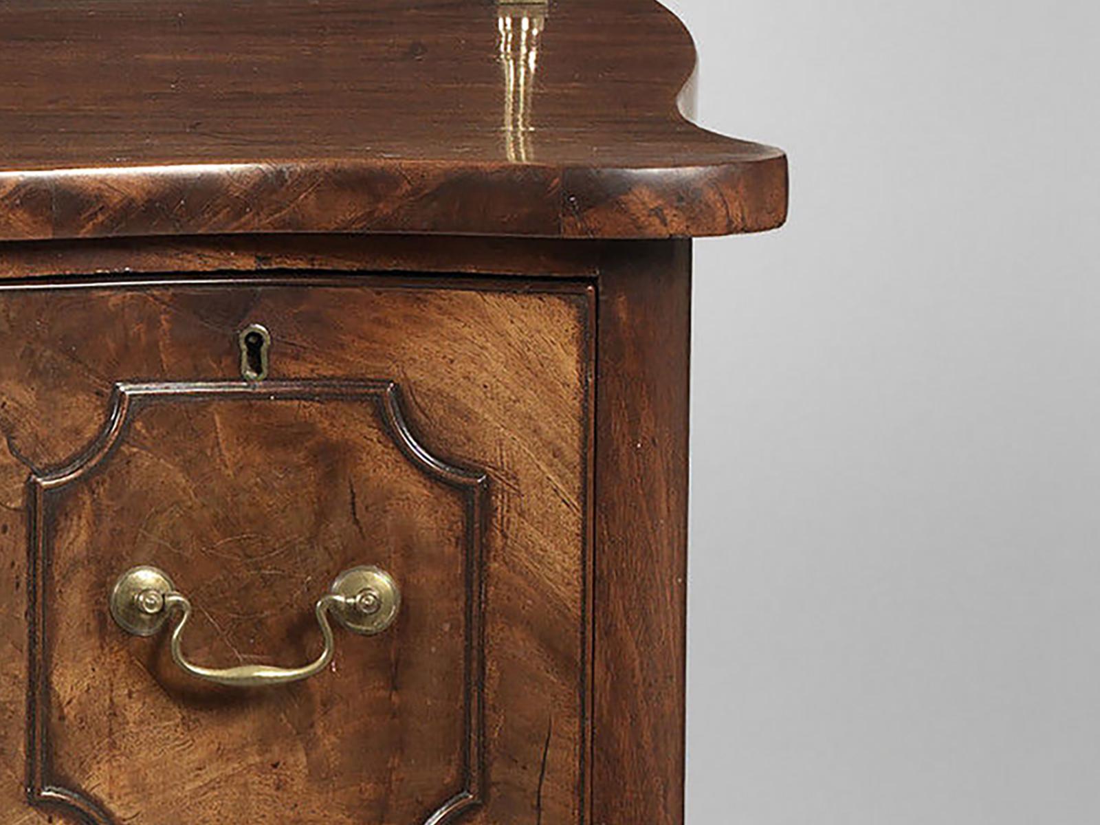 A George III mahogany serpentine fronted sideboard, the shaped top supporting two adjustable brass candle arms, above a central long drawer, flanked by twin drawers; all having fielded panels with astragal mouldings on square legs.

The sideboard