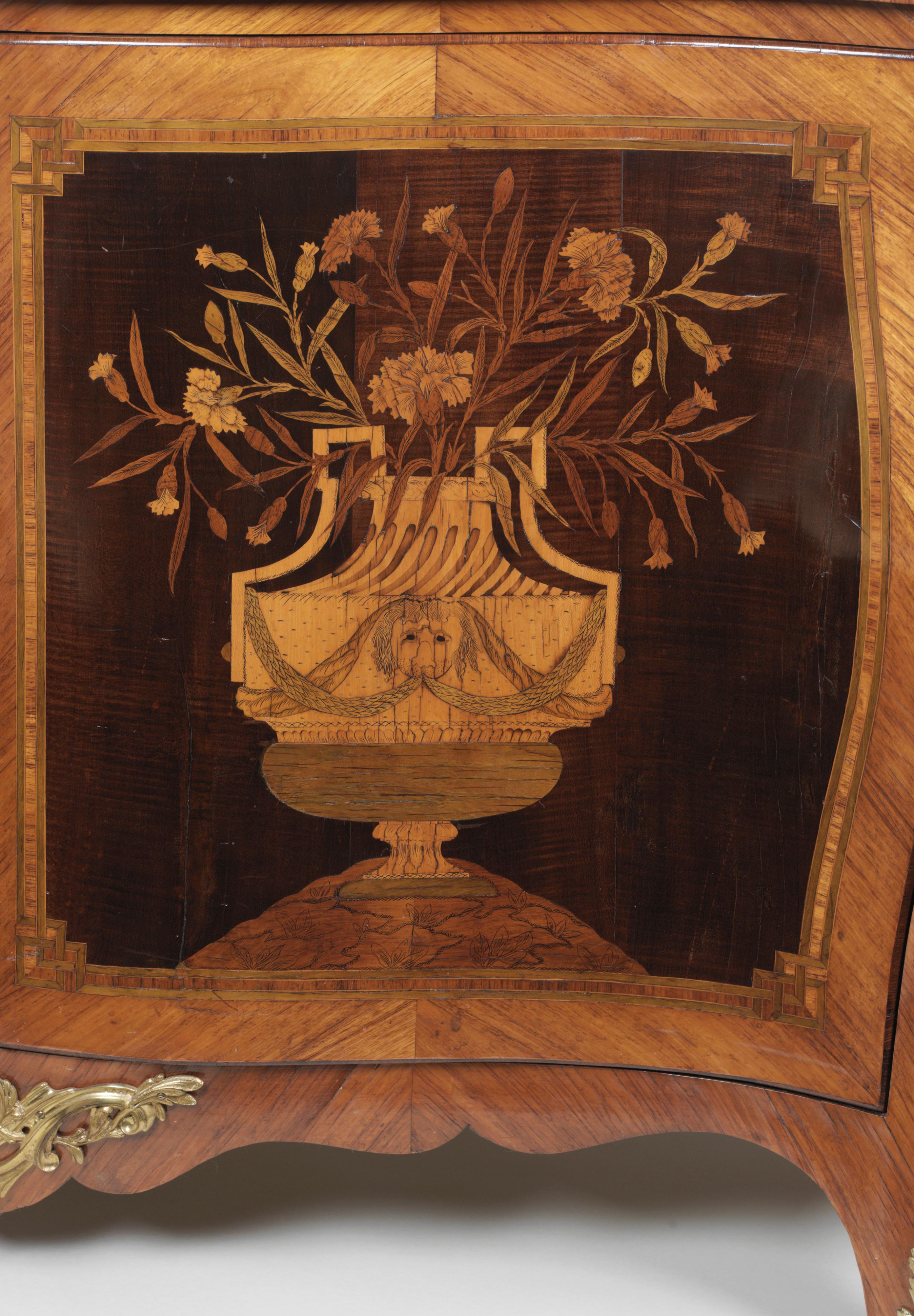 The top of the serpentine commode is inlaid with a marquetry panel of musical trophy hanging from a ribbon within a foliate cartouche, flanked by sprays of ribbon-tied flowers, on a background of harewood. It has a broad cross-banded border and