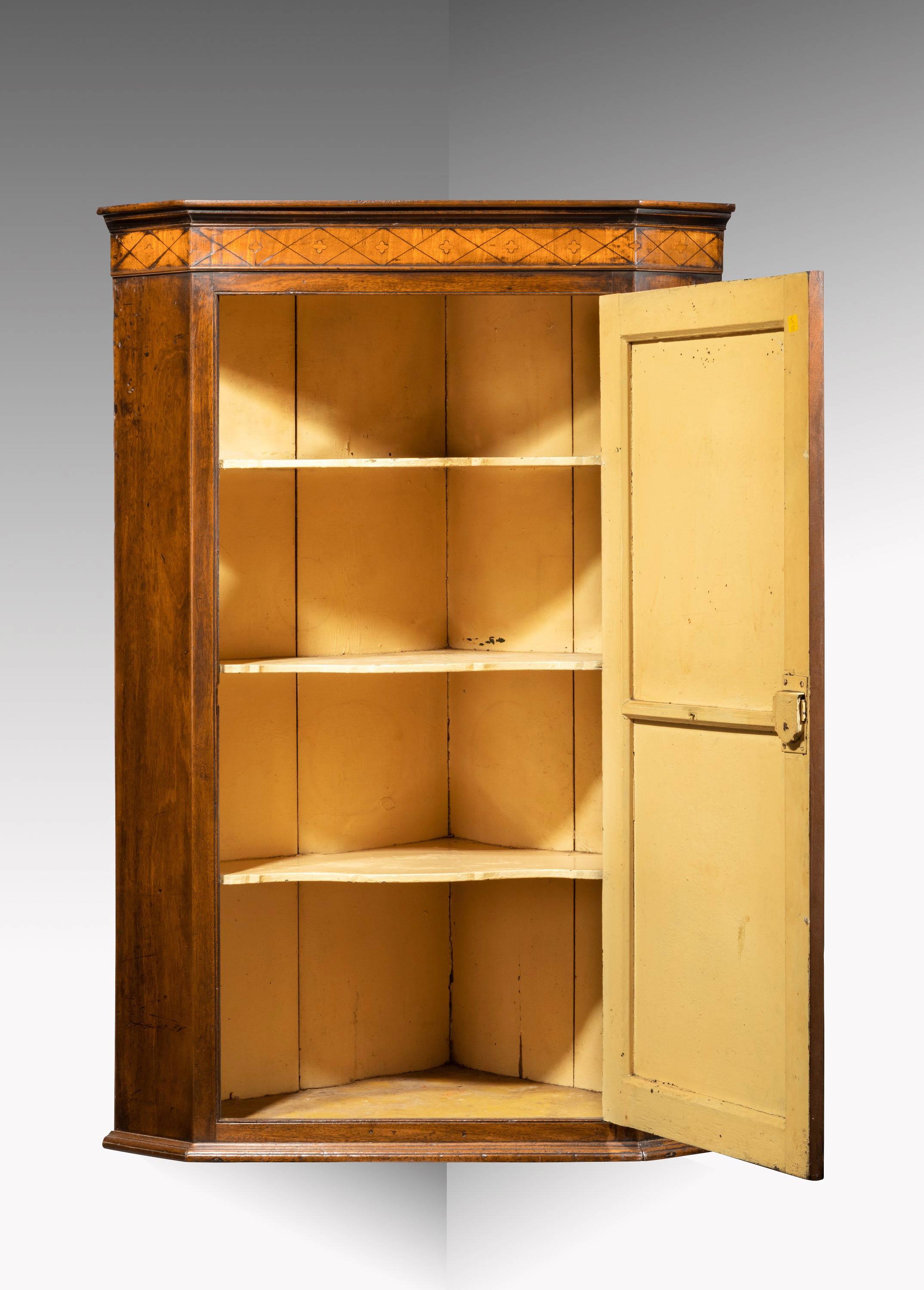 A George III mahogany corner cupboard. The drop frieze in boxwood and satinwood with parquetry decoration. The centre door with a well carved and inlaid shell. Showing signs of age and patina. The interior retaining the original serpentine