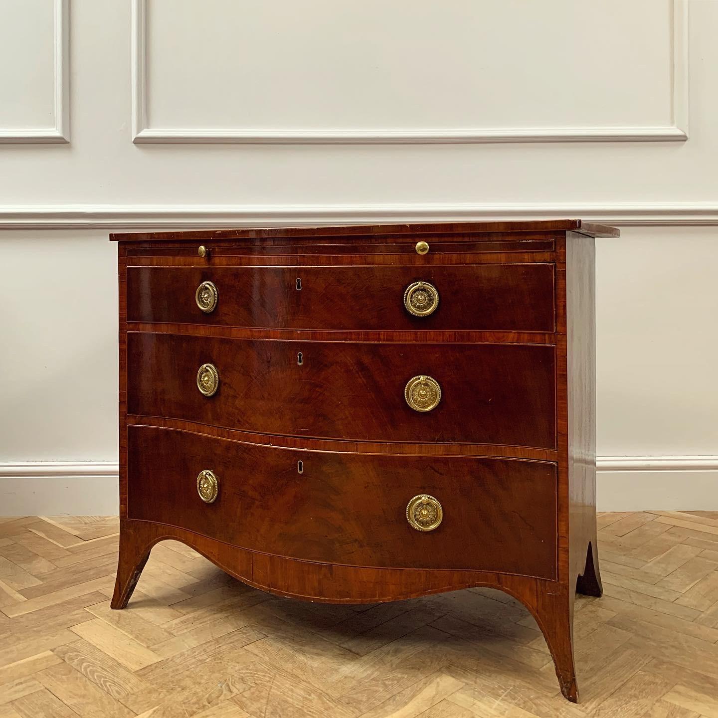 An elegant serpentine fronted commode in well figured mahogany bordered with box and rosewood above shaped brushing slide with original baize above three graduated drawers, original gilt brass ring pulls, shaped apron and swept bracket