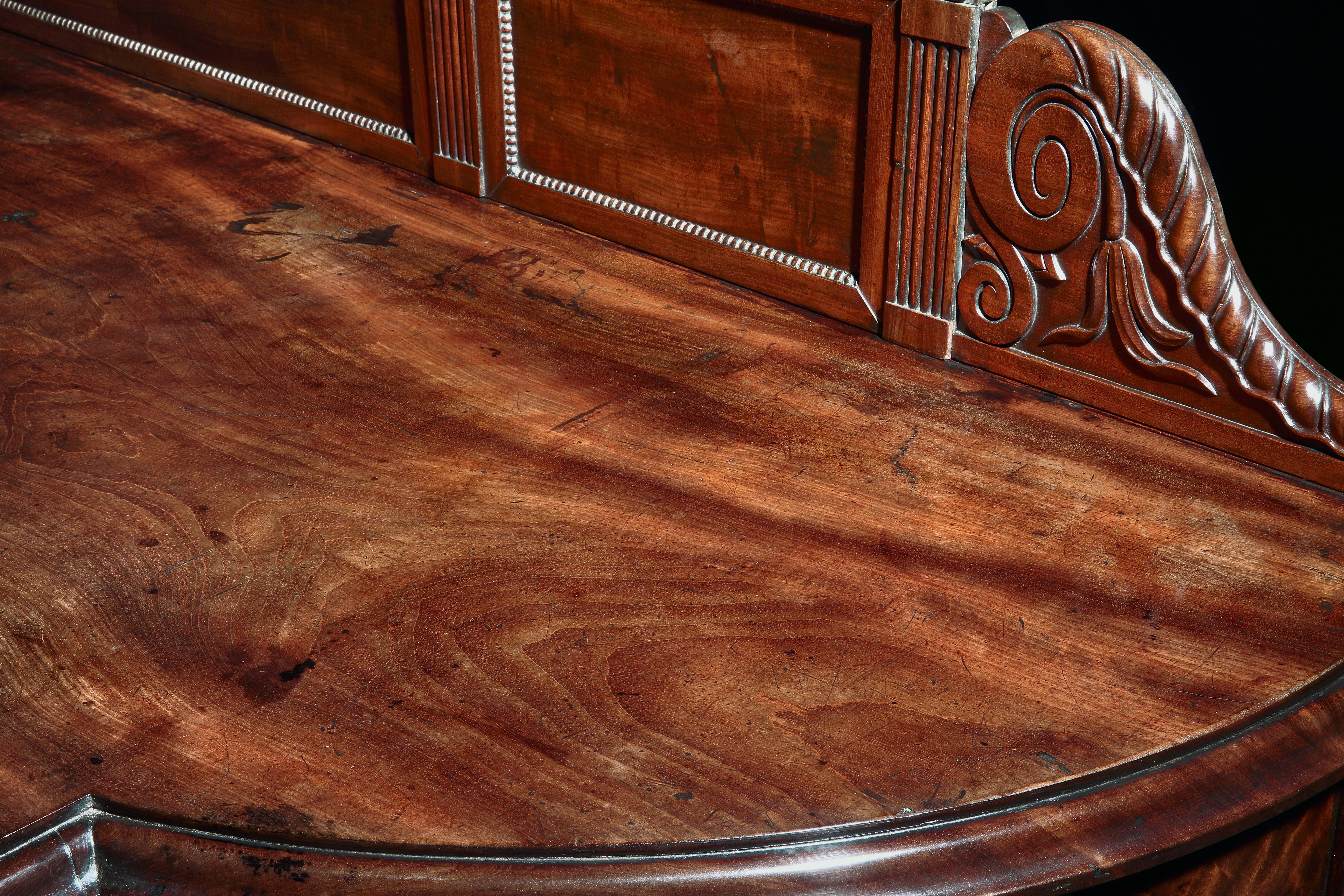 A monumental George IV Irish brown mahogany serving table. The beautifully figured inverted breakfront top with bow ends and moulded edge, above a panelled frieze, resting on six turned and fluted acanthus carved legs, with a three panelled splash