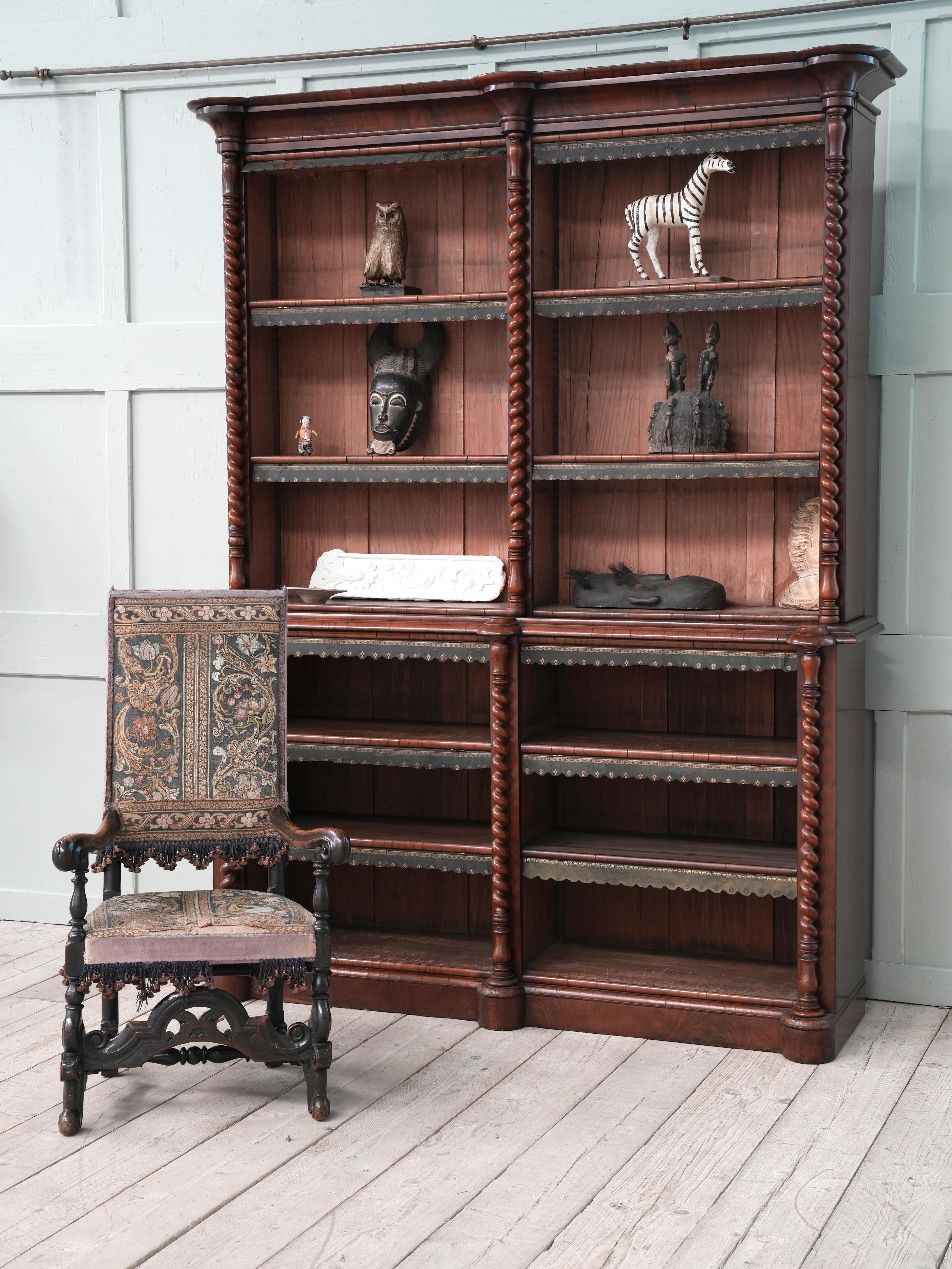 The shaped cornice above two open bays of adjustable shelves flanked and centred by barley twist turned uprights, the base has two adjustable shelves per bay, hinged timber dust covers with tooled leather coverings, raised on a shaped plinth base.