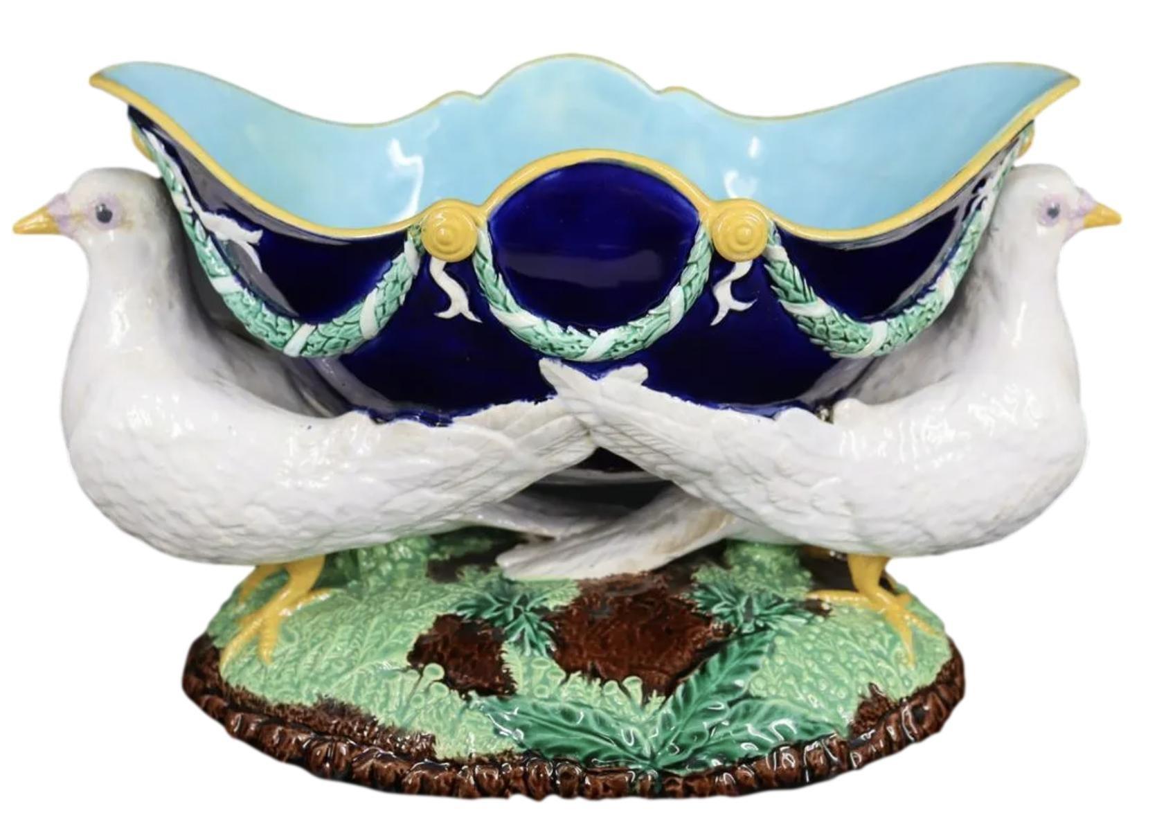 A George Jones Majolica Centerpiece Jardinière, the cobalt glazed shaped bowl supported by two naturalistically molded and glazed doves, with ribbon-wrapped garlands and trimmed in yellow ocher, on a rustic mound with green-glazed leaves and ferns