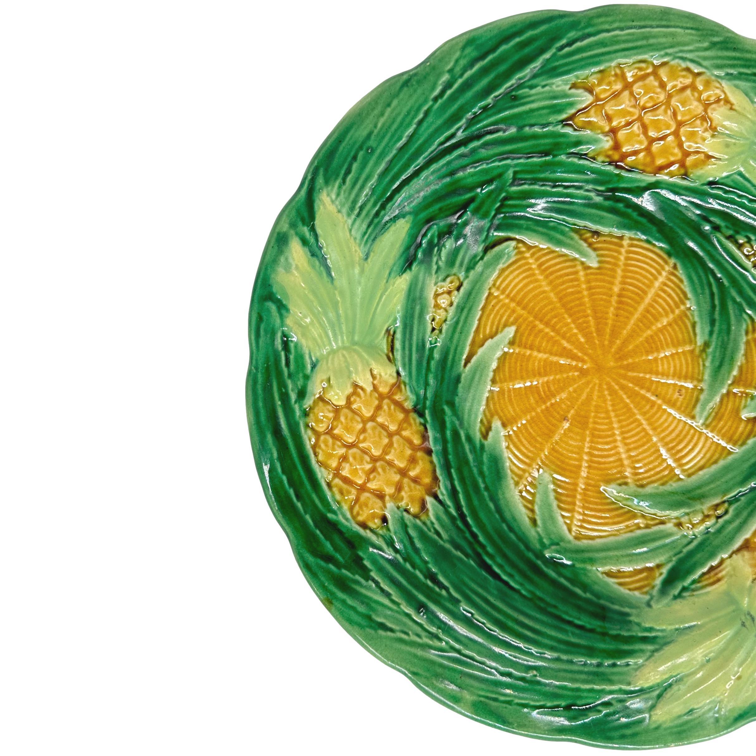 Assiette à dessert en majolique de George Jones, avec des ananas moulés en relief et des feuilles tourbillonnantes émaillées en vert, le centre avec une simulation de vannerie émaillée ocre jaune, le revers avec des marbrures typiques d'écaille de