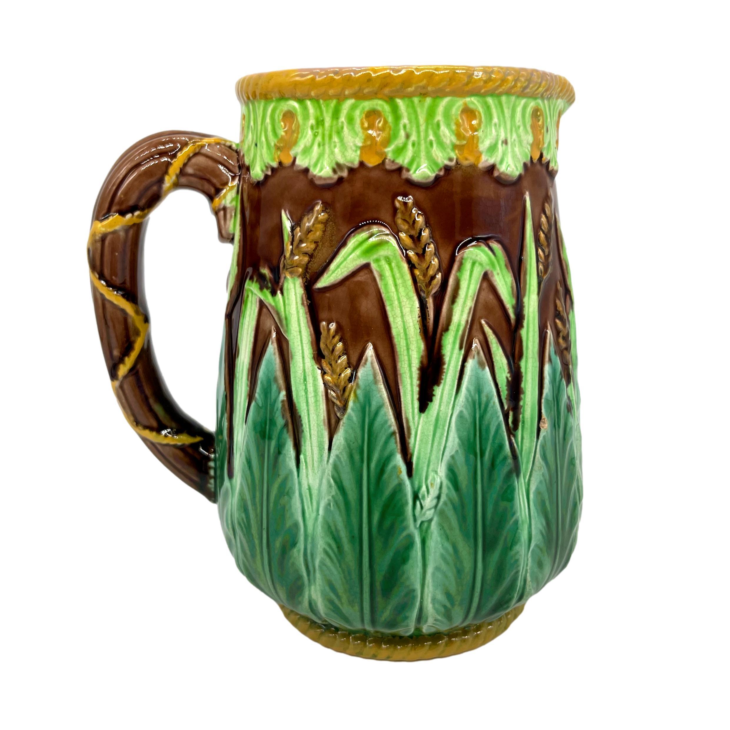 George Jones Majolica Pitcher, of baluster form with relief molded stiff acanthus leaves, grasses, and wheat on a brown ground, the top banded with stylized leaves glazed in chartreuse, with yellow glazed roping to the top rim and foot rim, the
