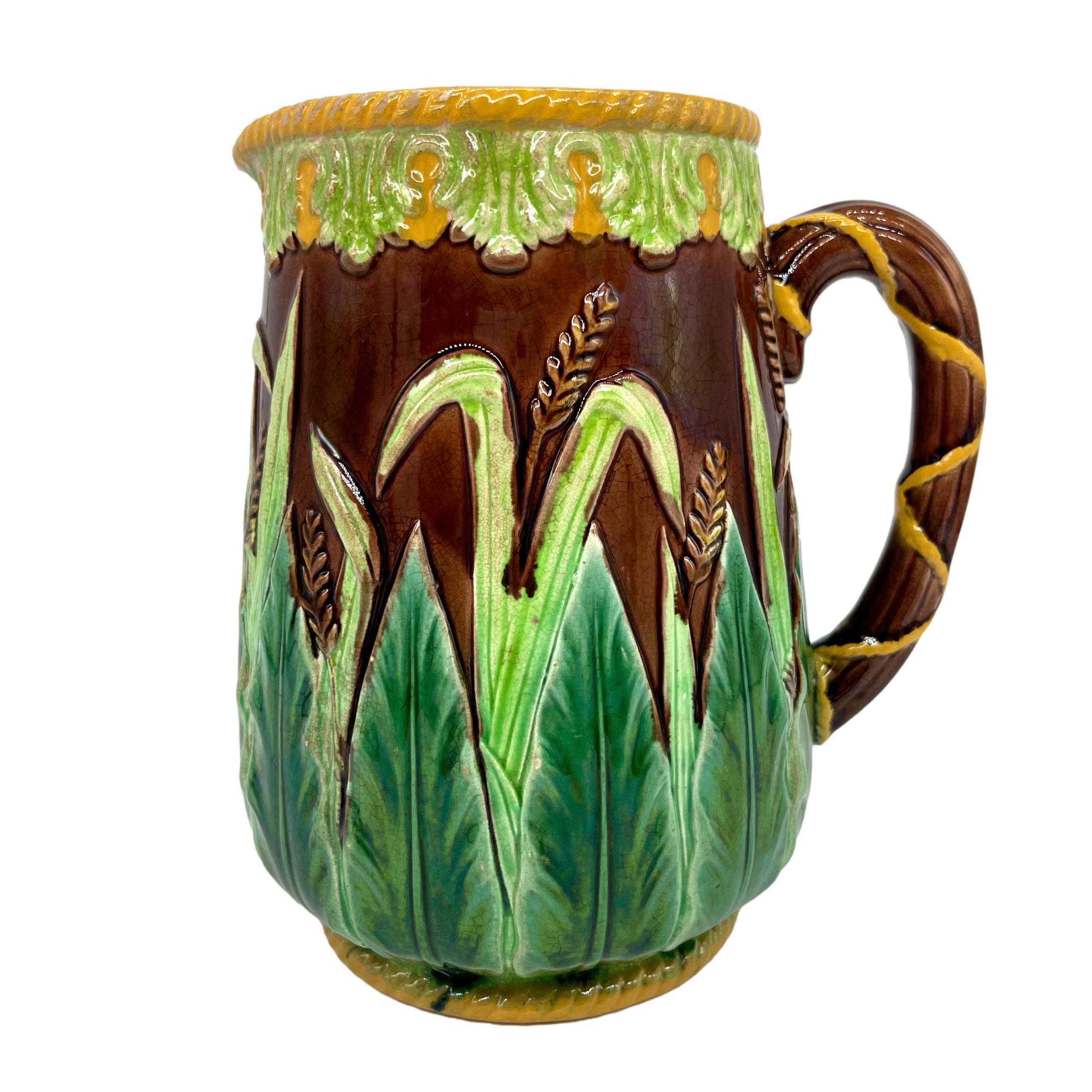 George Jones Majolica Pitcher, of baluster form with relief molded stiff acanthus leaves, grasses, and wheat on a brown ground, the top banded with stylized leaves glazed in chartreuse, with yellow glazed roping to the top rim and foot rim, the