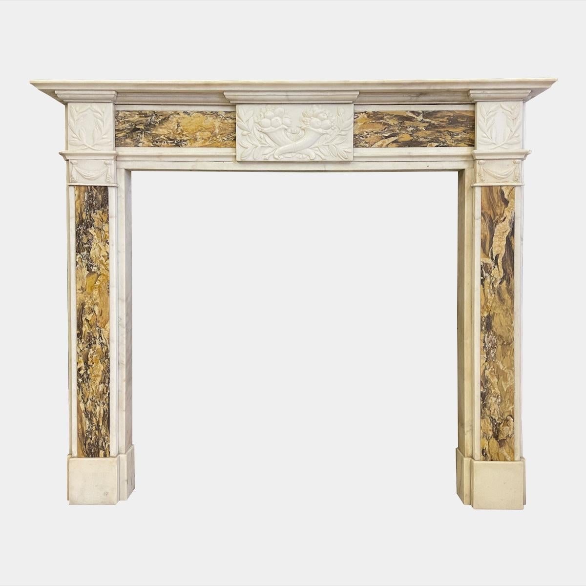 An early 20th century marble fireplace in the Georgian Neoclassical manner. Executed in Statuary white marble with Sienna Marble panels to the Jambs and flanking the frieze. The mantel being stepped and moulded, above the frieze with a carved centre