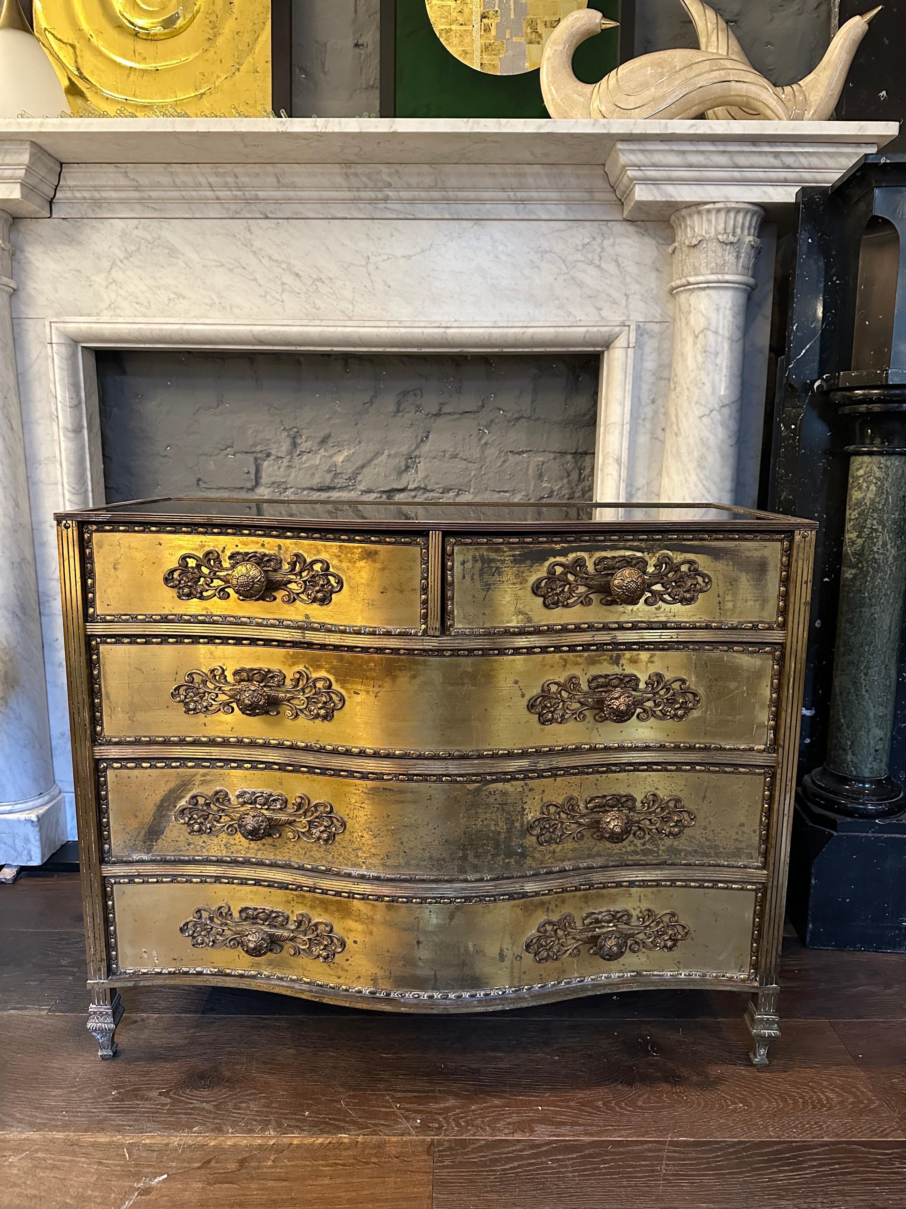 A gilt brass and black glass commode from the mid 20th century with gilt and now from age a rich worn patina bank of serpentine drawers. Having foliate scrolled mounts and decorative rosette handles. The reeded edges with black glass with to top and