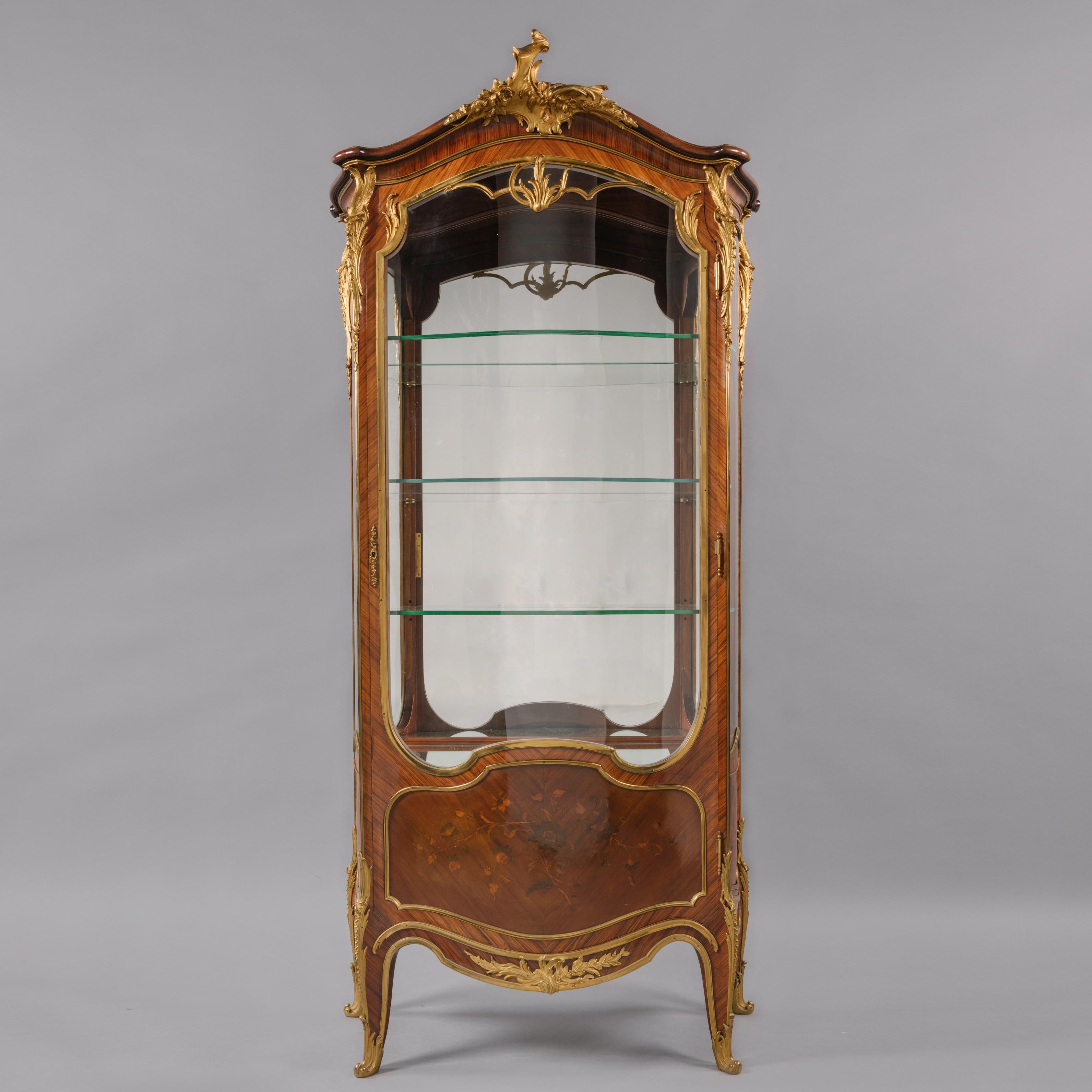 Stenciled to the back ‘Cruyen Paris, Ameublements de style’.

The arched top is centred by a gilt-bronze foliate mount above a shaped glass door to the front, opening to three adjustable glass shelves, with corresponding glazed side panels. Below