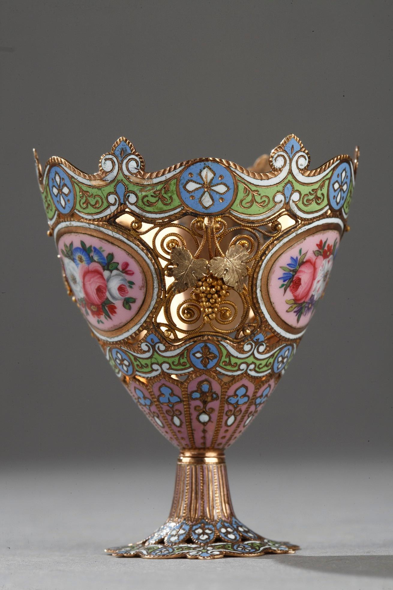 A gold and enamel zarf, pierced and enamelled. The collar is decorated with geometric patterns enamelled green and blue. The zarf has two medallions of pink enamel enhanced with floral motifs. The foot takes the geometric patterns of the