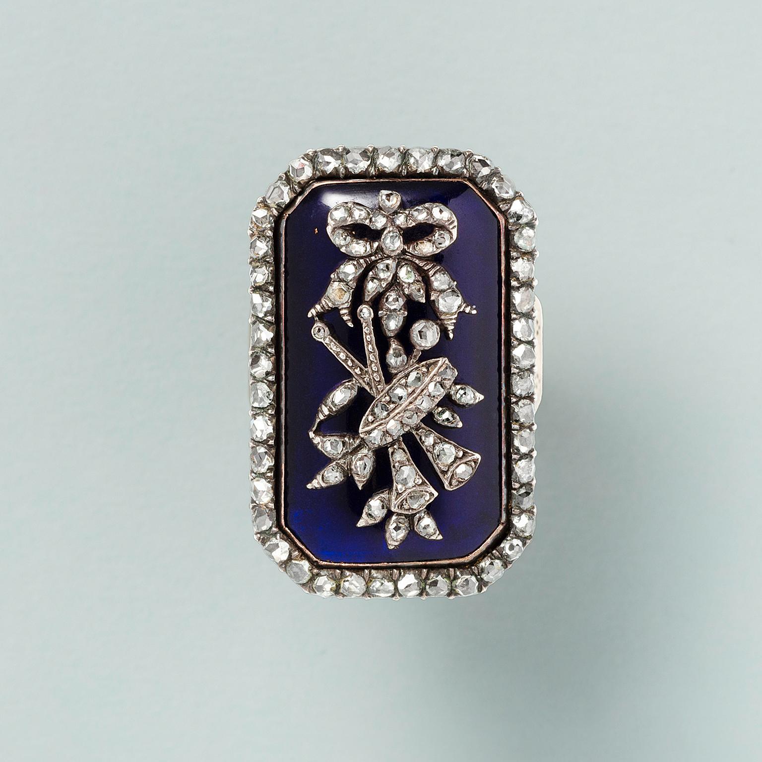 A gold and silver ring with a demountable octagonal panel set with an row of rose cut diamonds around a transparant blue glass panel with an silver ornament set with rose cut diamonds featuring  a bow followed and musical intruments and leaves. 18th