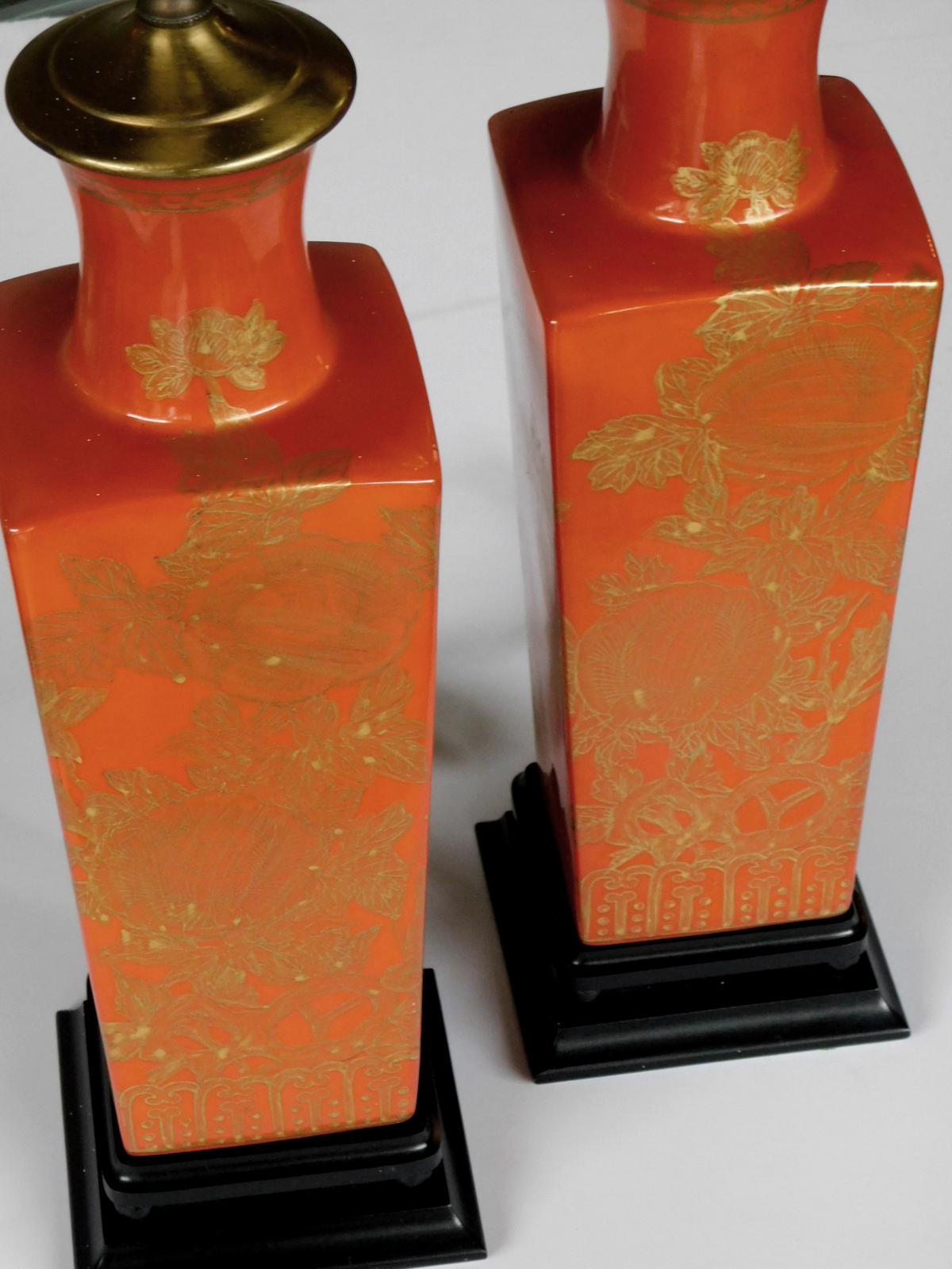 Each with flaring neck above square-form tapering body all in a richly-colored red-orange glaze decorated with subtle gilt floral vines; resting on a wooden ebonized based