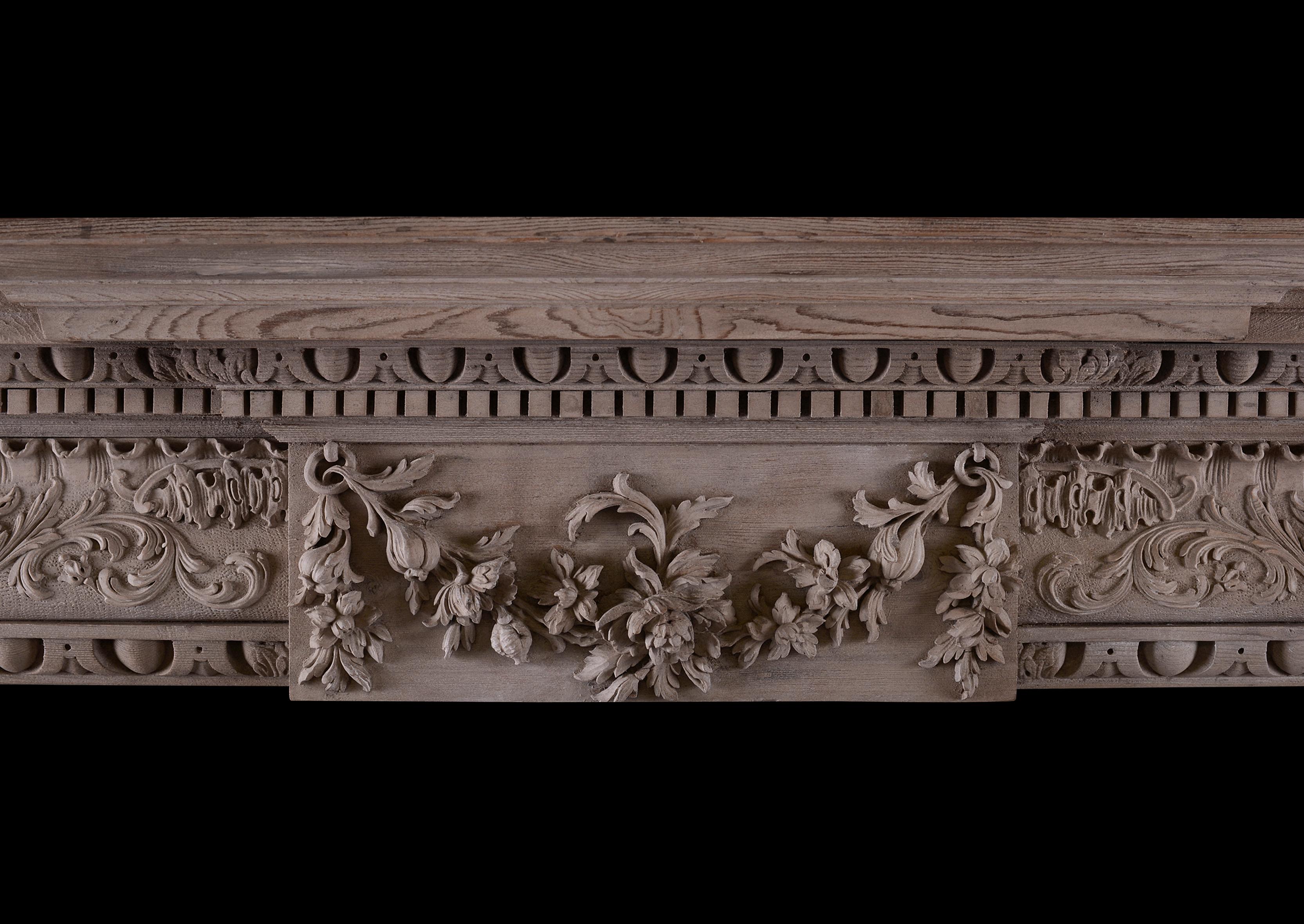 A carved bleached pine fireplace in the Georgian manner. The break-leg jambs carved with egg-and-tongue moulding and ornate decorative scroll work. The frieze with ogee moulded frieze with carved scrolls and foliage throughout, the centre blocking