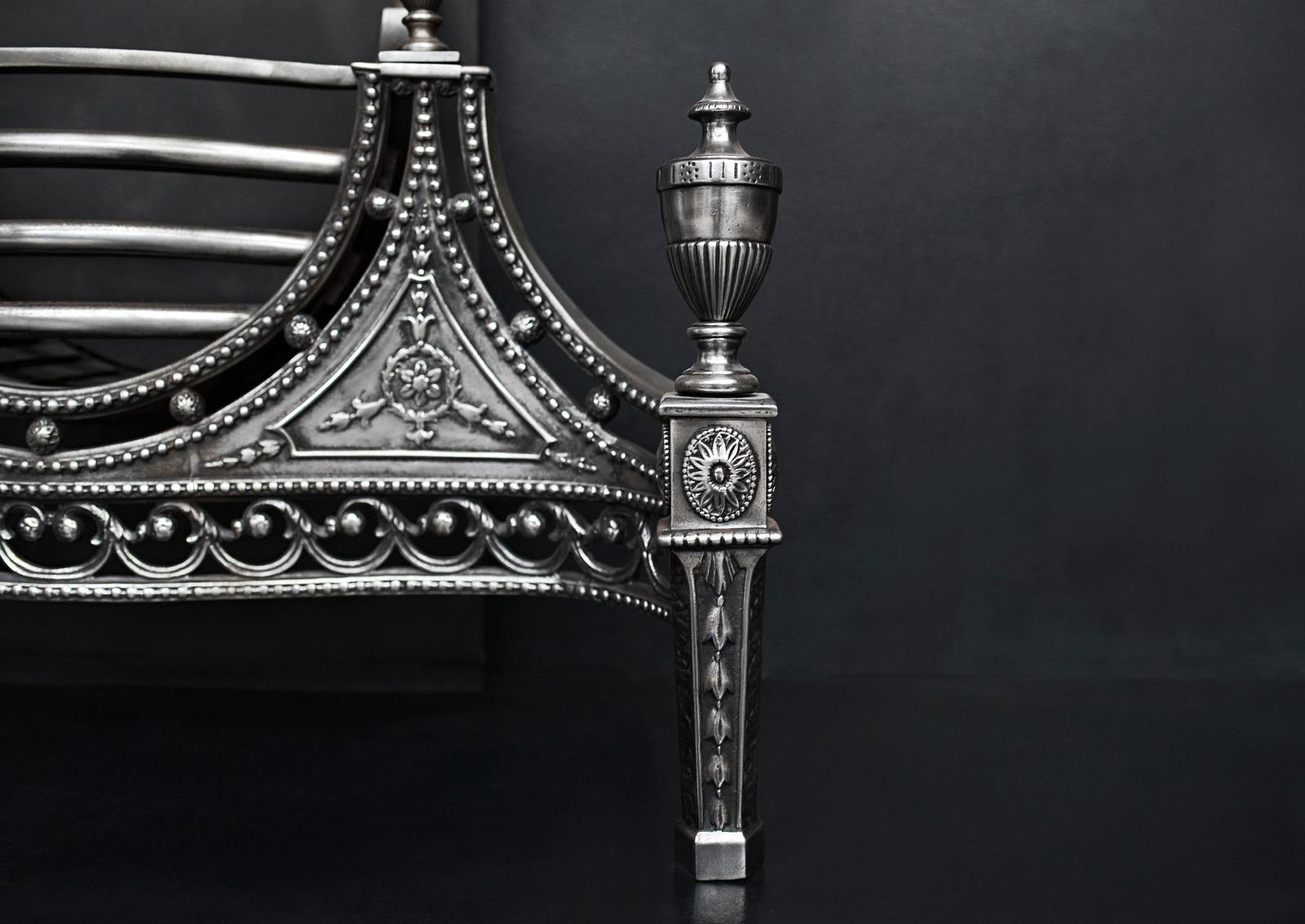 An English cast iron fire grate in the Georgian manner. The burning area grate surmounted by a semi-circular plate embossed with a radiating dart and bellflower design, serpentine-shaped scrolled and pierced fret surmounted by urn finials. The