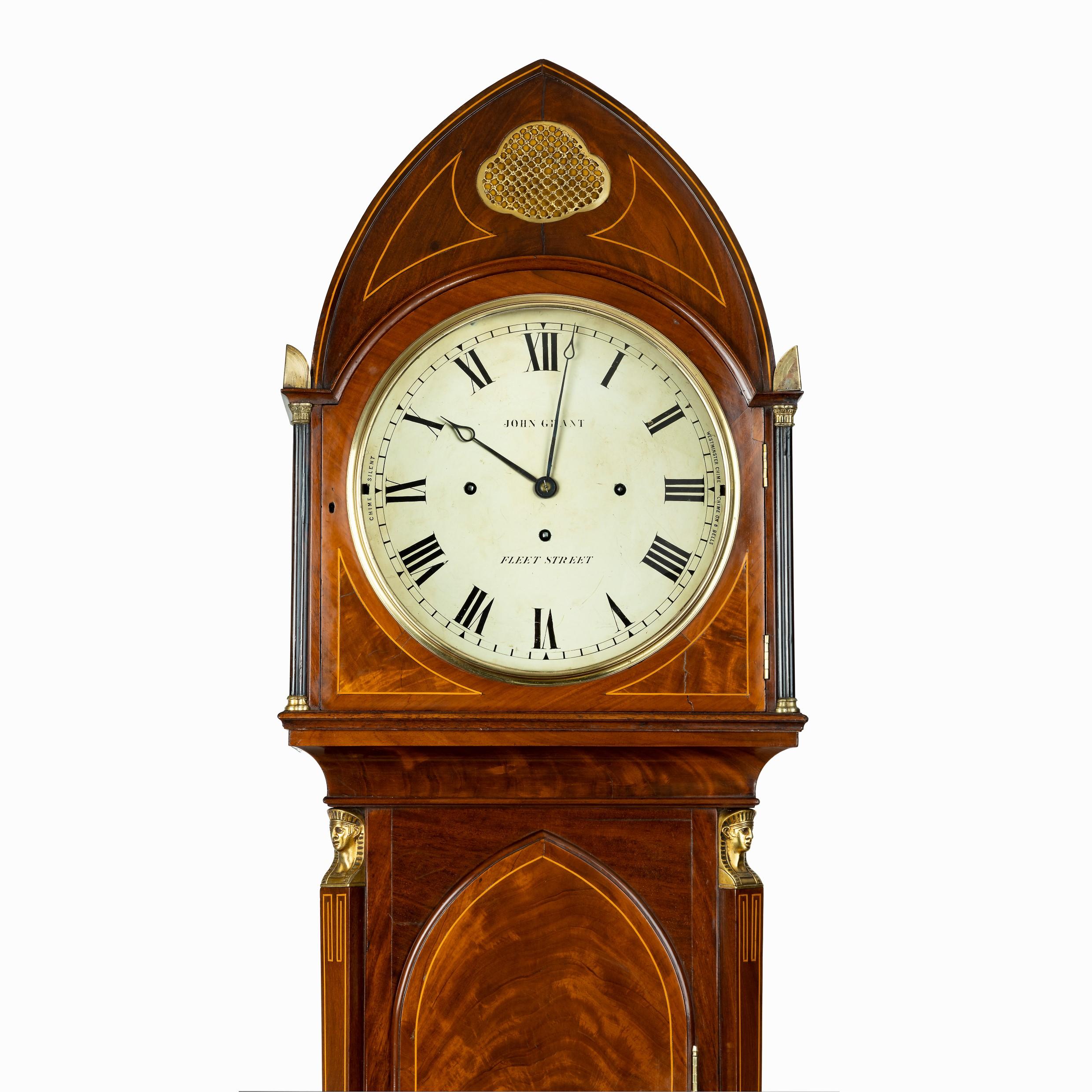 A good quality Regency ‘Egyptian style’ mahogany longcase clock by John Grant, the case surmounted by Egyptian columns with heads and feet of cast gilt brass, the lancet style hood with an open brass fret above the dial and on the sides, backed with