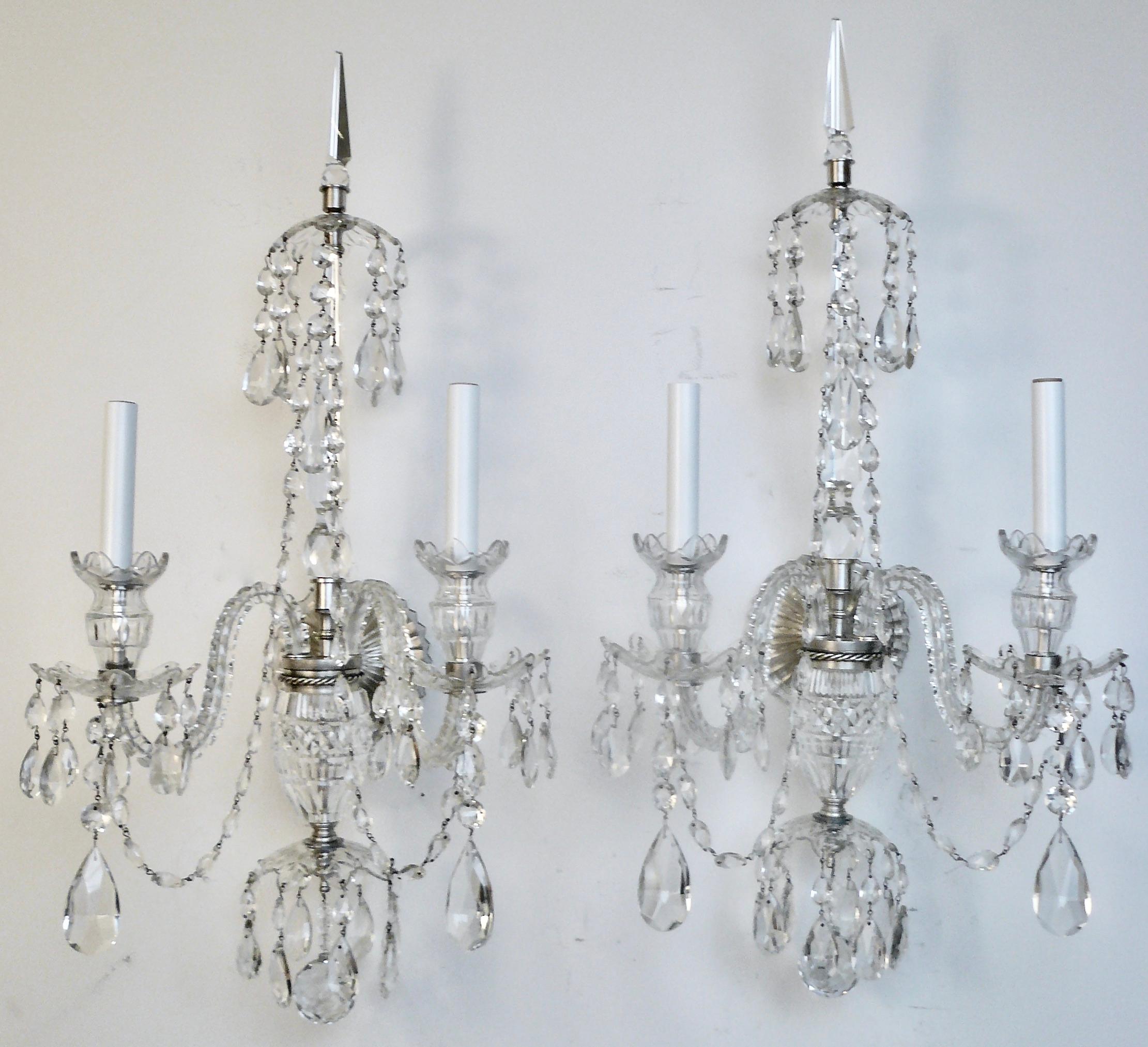 This large scale pair of Georgian style cut crystal two light sconces feature crystal spikes, swagged chains of graduated almond shaped prisms, and and silvered fittings. The sconces have faceted ball finials, Van Dyke style bobeches, and terminate