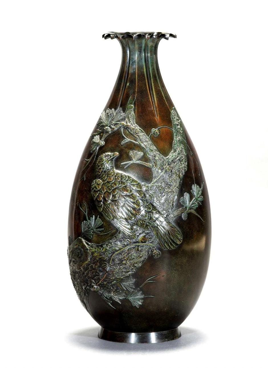 Große Bronzevase mit grünlich-rötlicher Patina, die einen Falken mit nach rechts gewandtem Kopf auf einem großen Ast darstellt, der die Vase vom Boden bis zum oberen Hals schräg umschlingt. Lappiger Rand, von dem einige Rillen ausgehen und sich