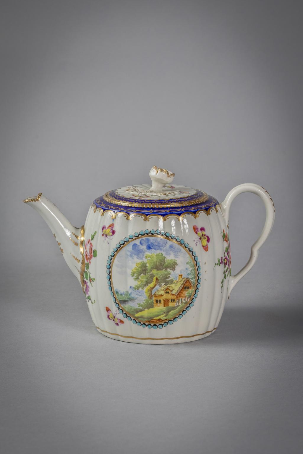 Each painted with landscape roundels flanked by flower sprays within a narrow blue and gilt border, comprising: a fluted barrel-shaped teapot and cover and a hexagonal fluted stand; a covered sugar and another hexagonal fluted teapot stand, open