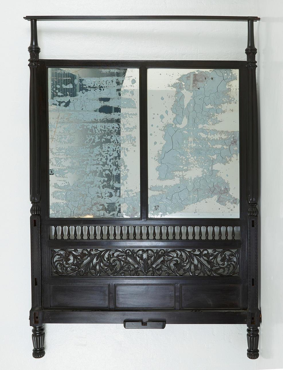 An queen-sized Colonial poster bed. The headboard with a double-paned mirror and intricately carved details. Mattress supported by low slats, a low-profile box spring is recommended. Circa 1900.