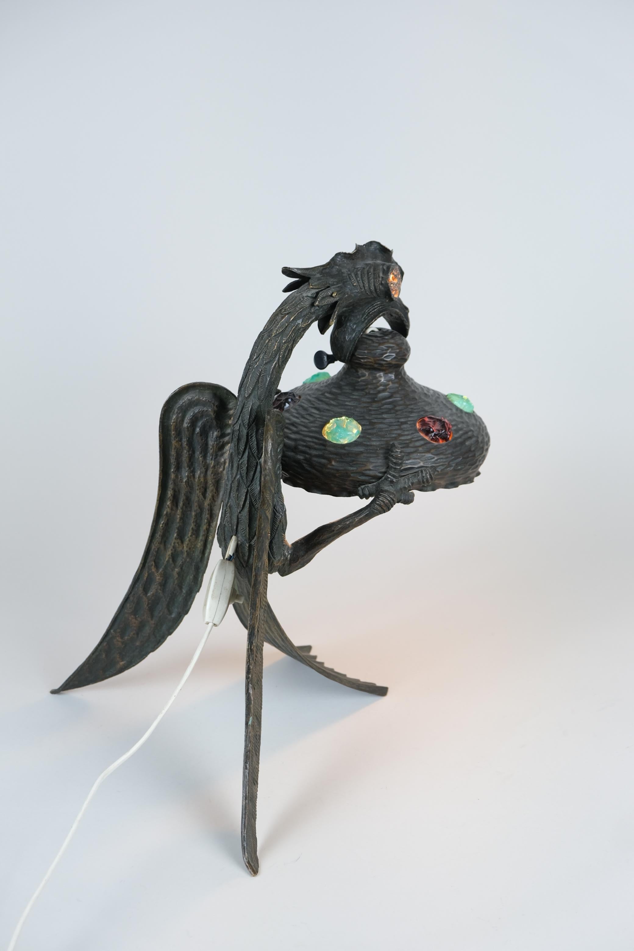 Une lampe de table exceptionnellement grande, faite à la main, martelée et coulée en bronze, avec de magnifiques décorations en verre chunk coloré. Cette lampe représente un aigle aux traits mythiques qui tient l'abat-jour avec son bec. La tête de