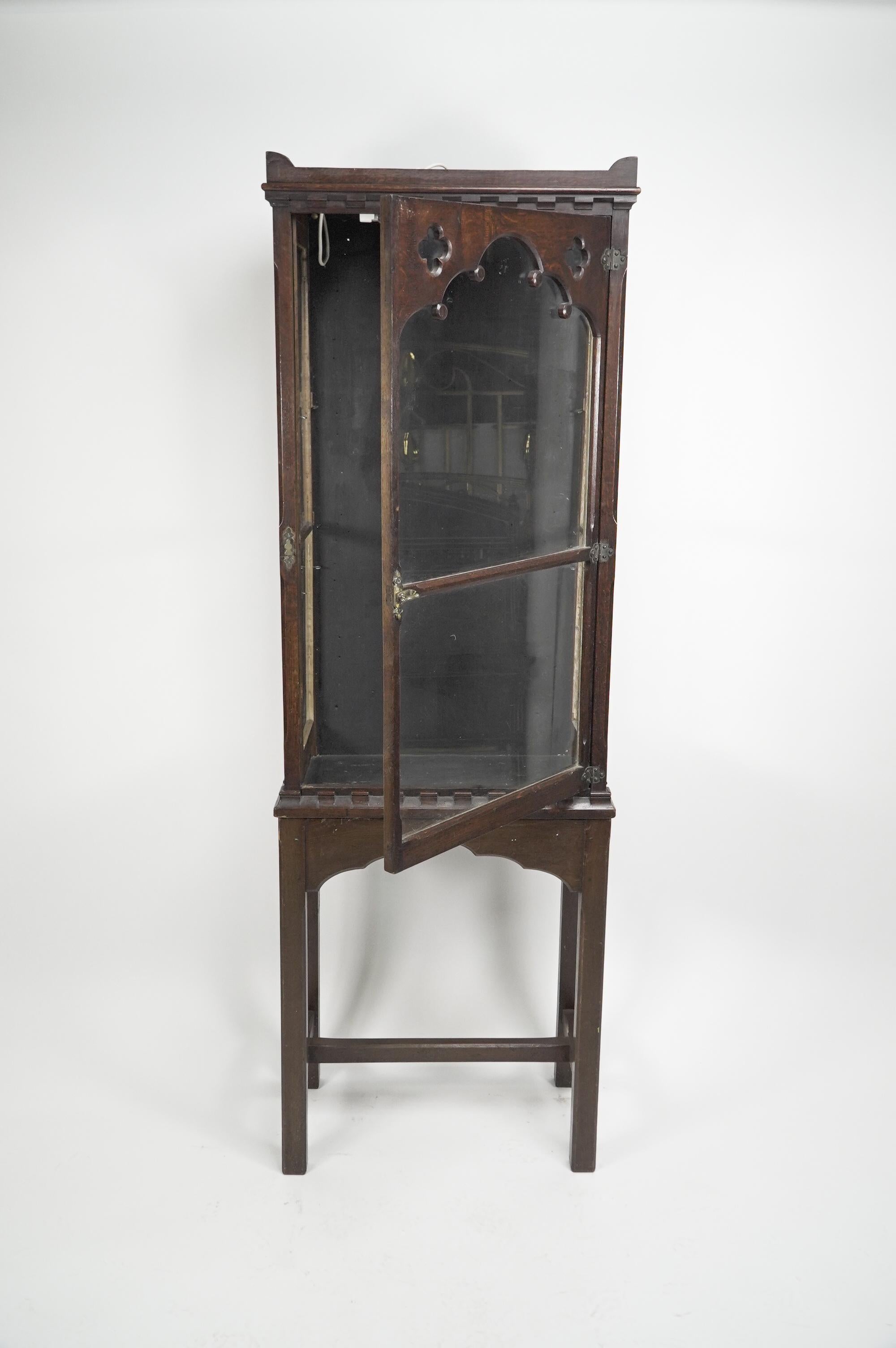A Gothic Revival oak display cabinet with an open display area to the top and little ear shaped details to each side and a castellated decoration below the glazed door, with five arches to the upper section and pierced quatrefoils to each side, and