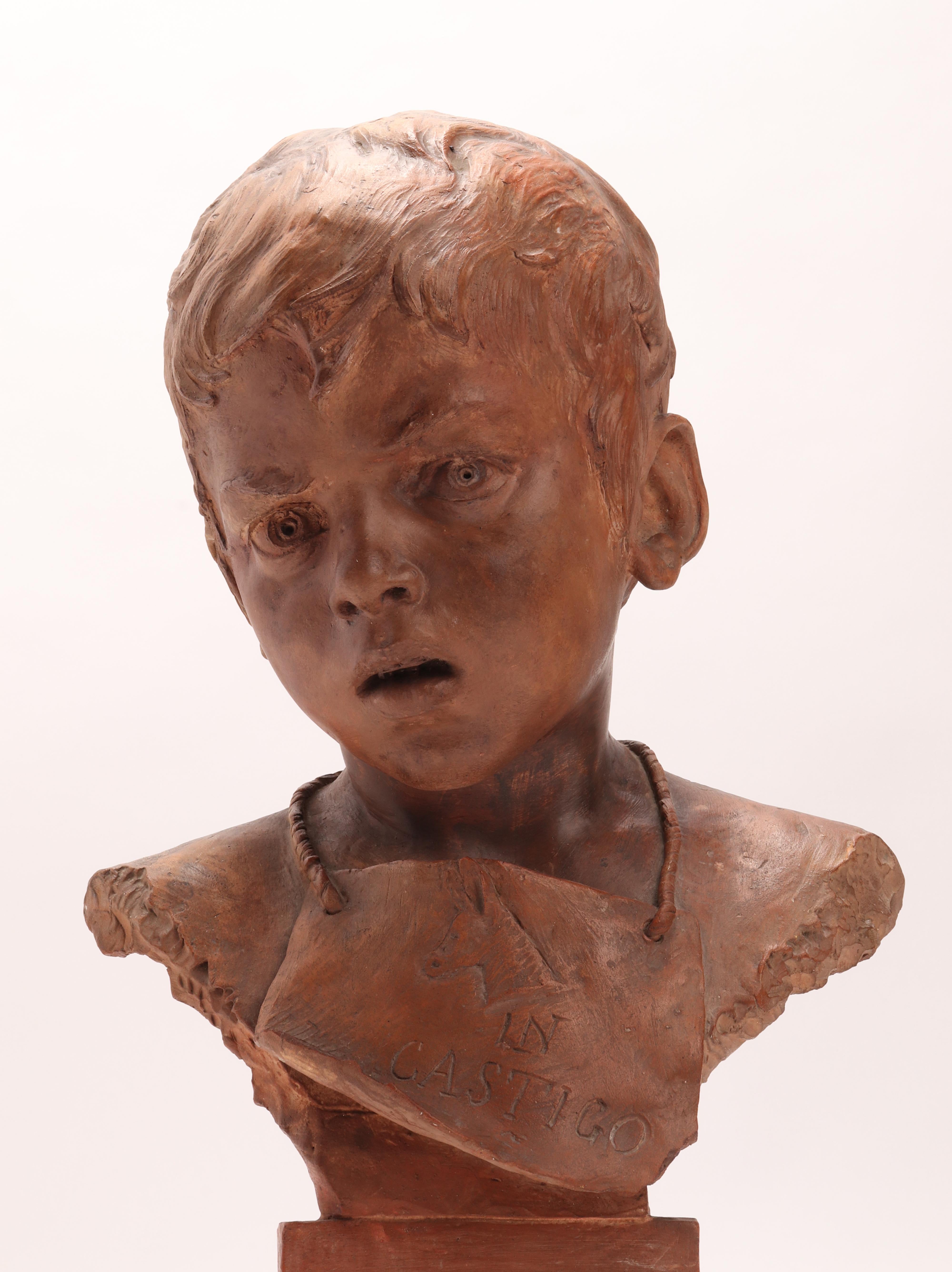 A handmade terracotta sculpture. It has a circular base with a wavy edge, then narrows to become a cylinder. On the base rests the bust of the child wearing an apron held around his neck by a lace. On the apron, a donkey's head is engraved in