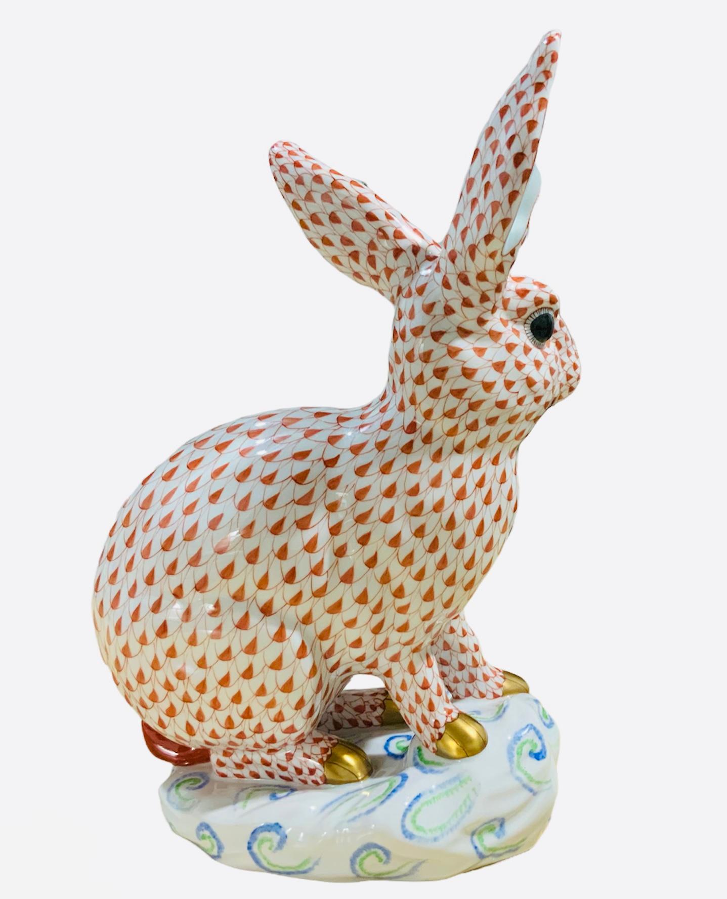 This is a Herend porcelain hand painted large rabbit that is sitting up above an asymmetrical oval white rock decorated with blue and green C-scrolls. Its background is white with orange fish scale net pattern. The inside of the ears are light green