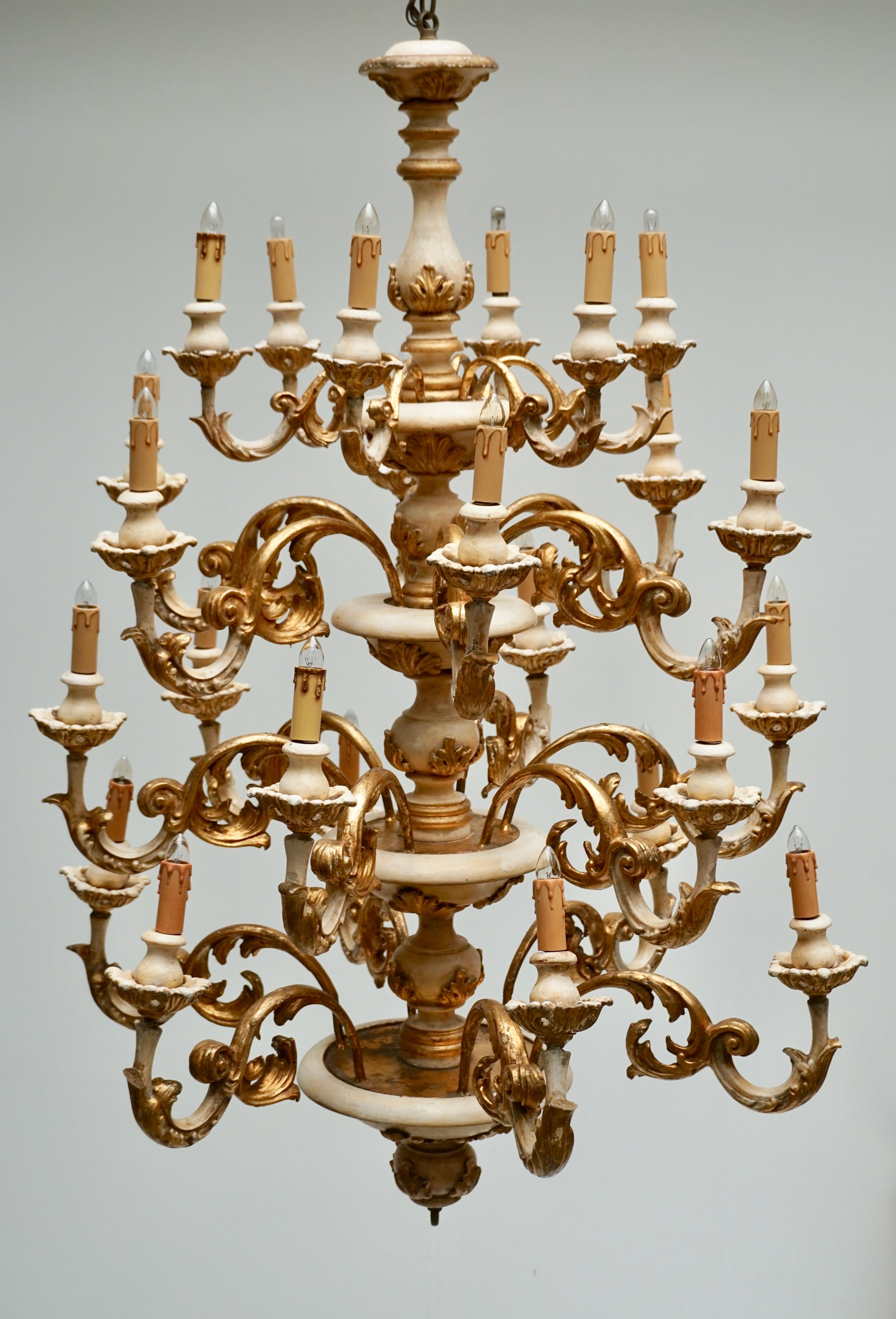 A highly decorative and elegant wood, metal and stucco 24–light electrified chandelier.
The central shaft with its four–level division is made out of cream–painted wood and leafy gilt stucco ornament, the four tapering rows of each six scrolled