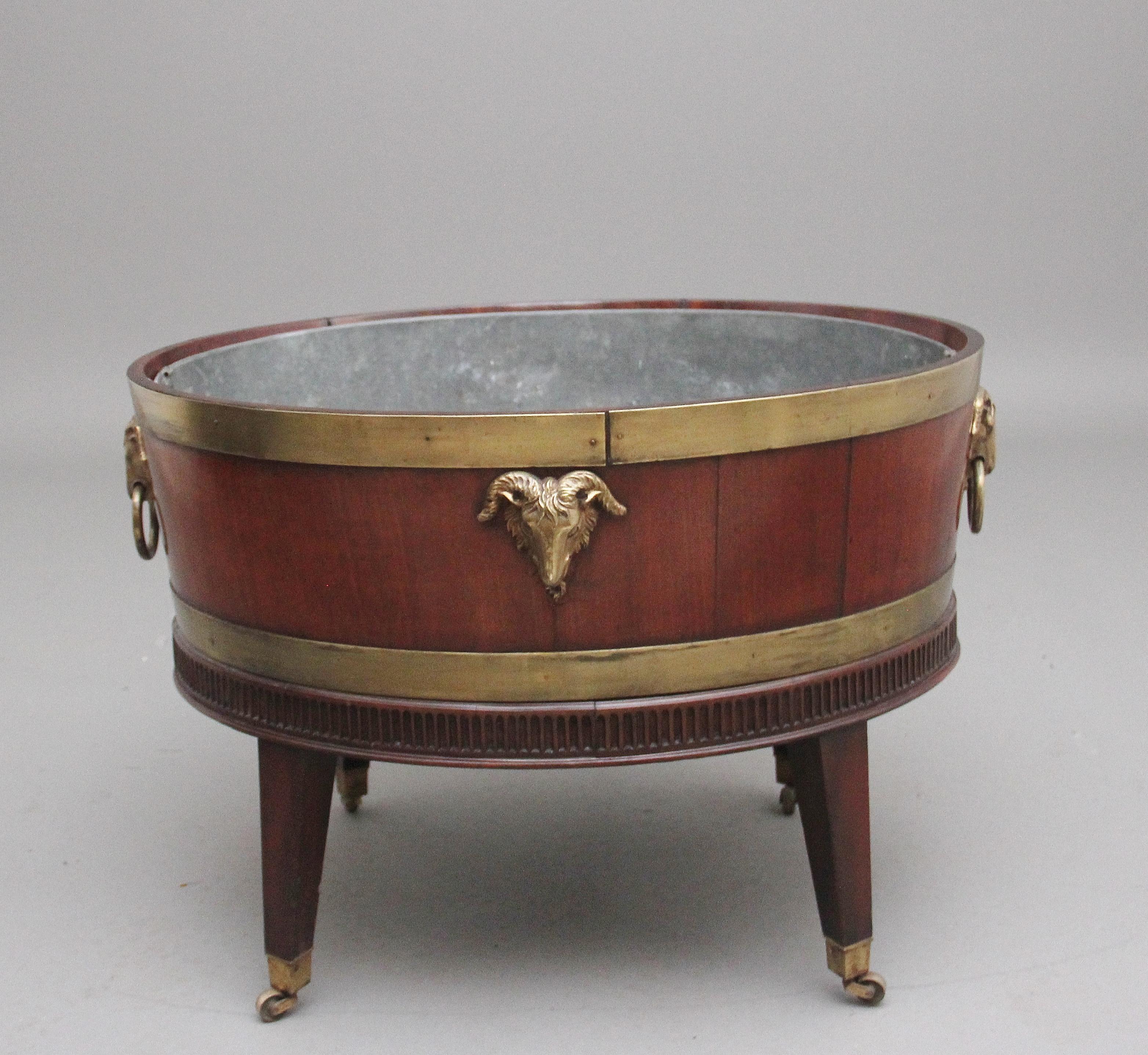 A highly decorative early 19th Century mahogany and brass bound wine cooler of an oval shape, having the original zinc lined interior for storage of various alcoholic beverages, having brass straps at the top and bottom, the sides and ends of the