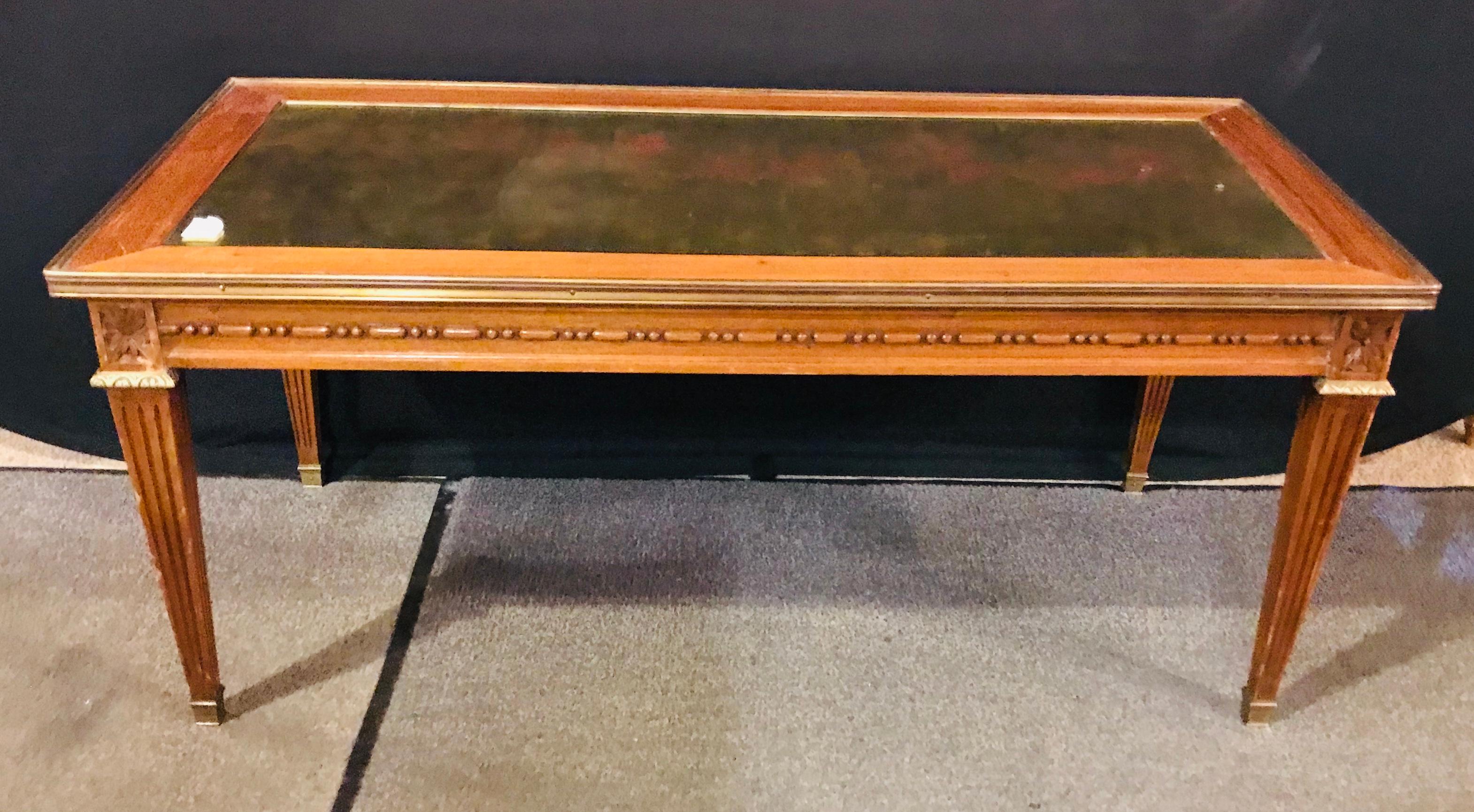 A mahogany gilt glass top Jansen style coffee table in the Hollywood Regency style with brass mounts throughout. Having tapering legs supporting a carved apron frame.